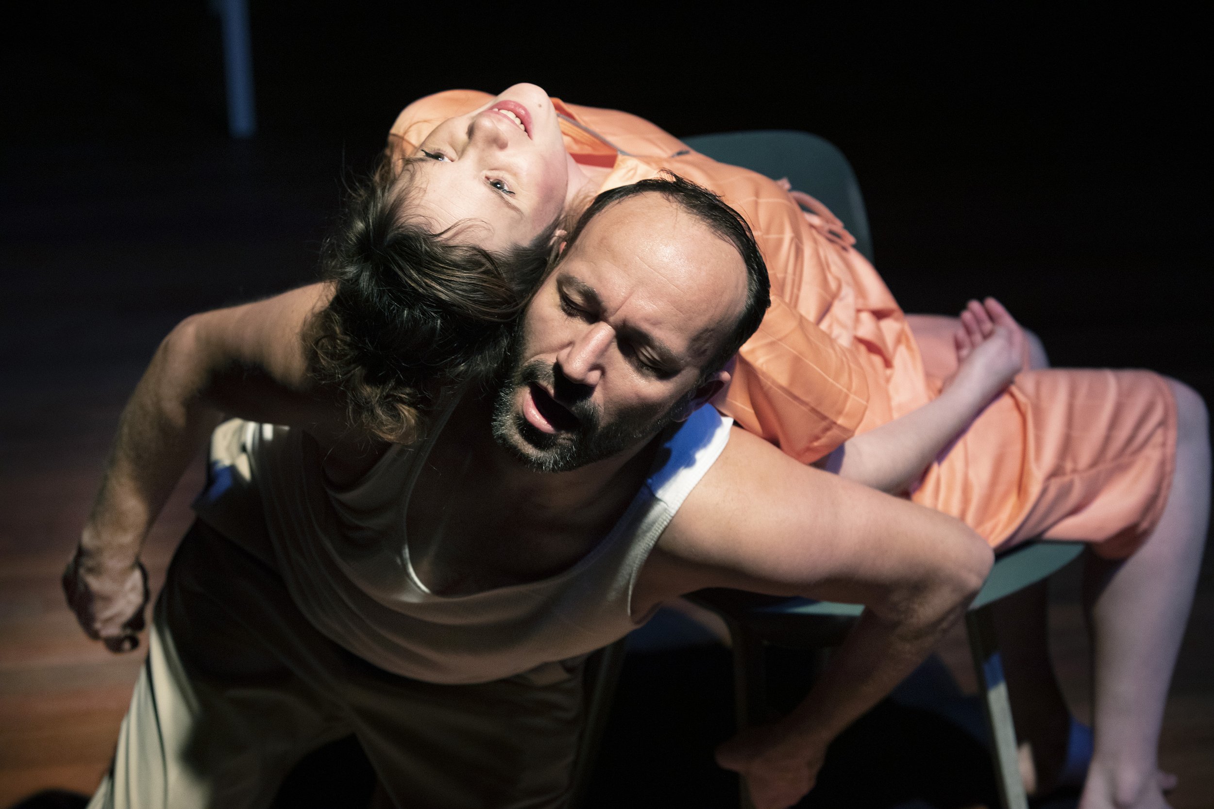 Two actors performing a theatrical scene, one man and one woman, with the woman leaning back over the man's shoulders.