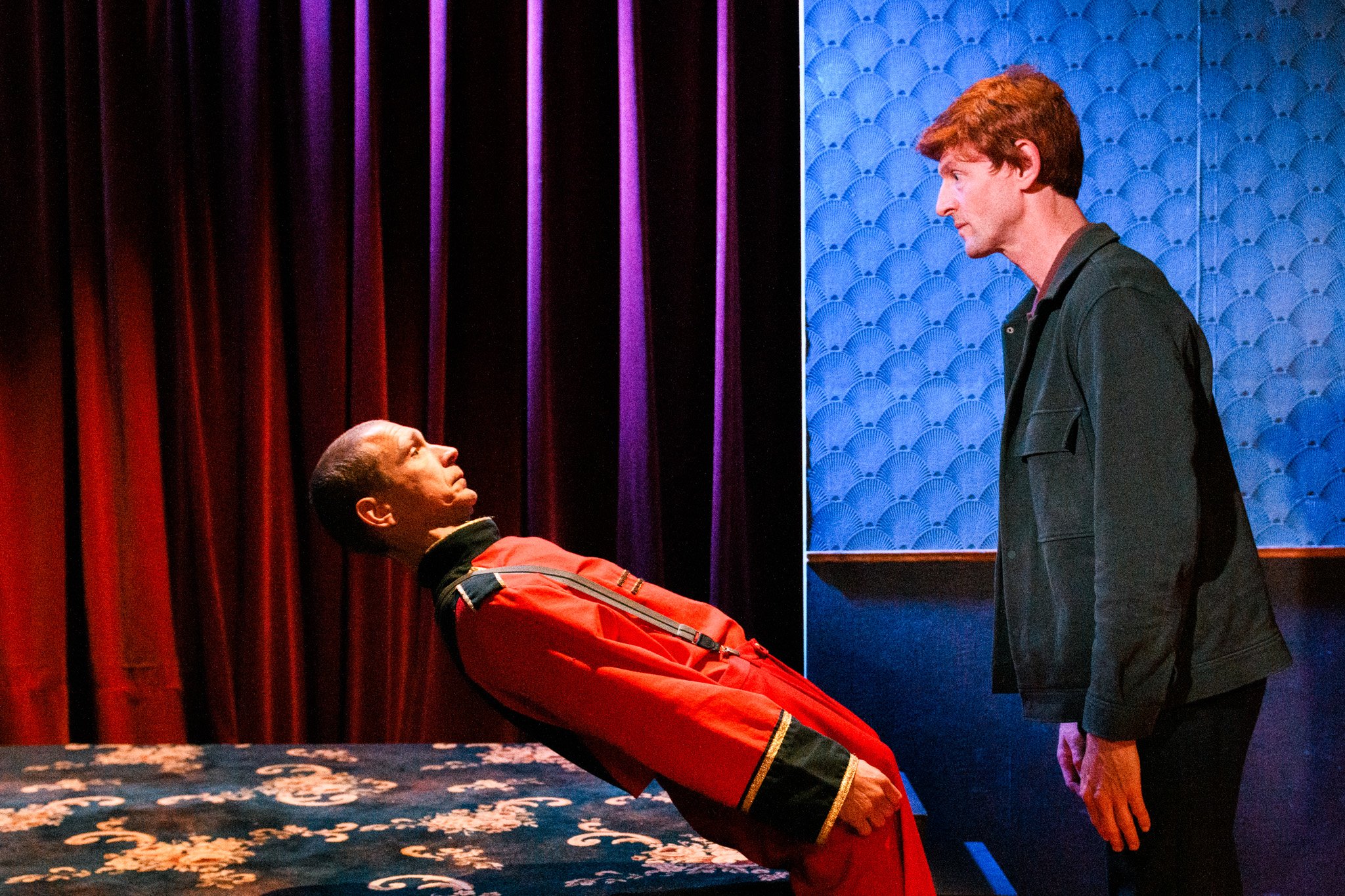 Two men facing each other in a dimly lit room with red curtains and blue textured walls, one leaning back on a table and the other standing.