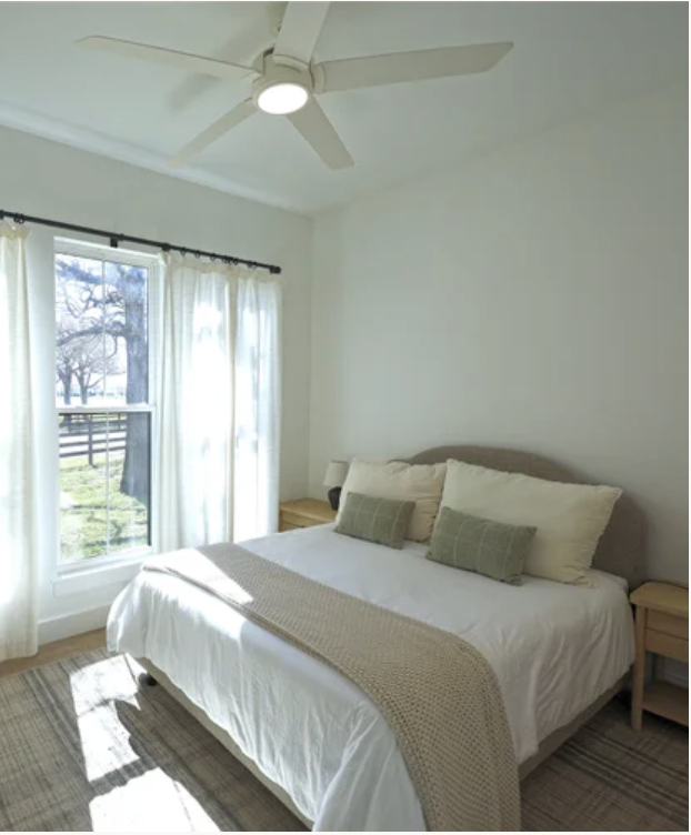 Bright bedroom with a white bed, green pillows, beige throw, wooden nightstands, window with white curtains, ceiling fan, and a view of trees outside.