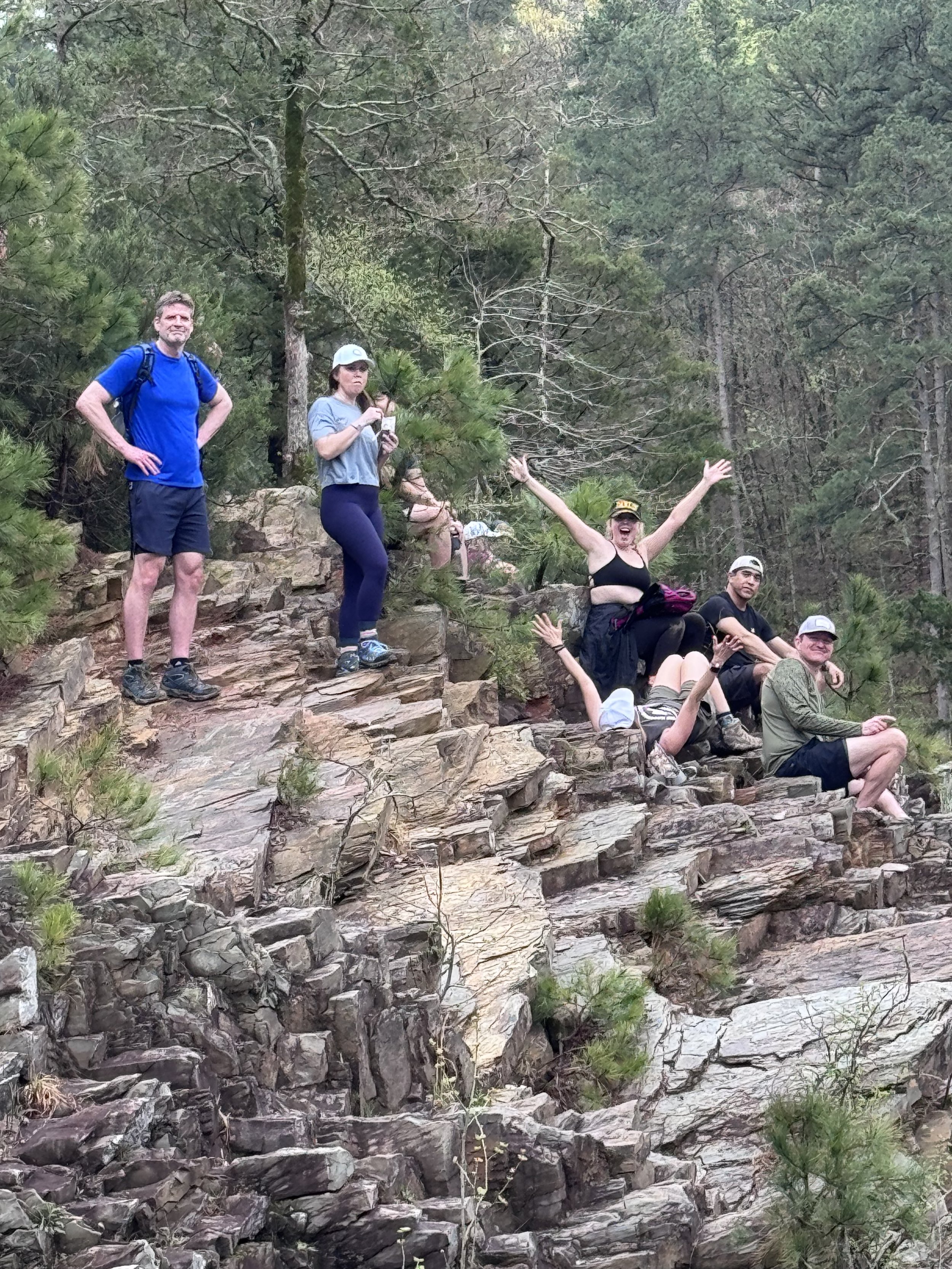 A group of people on rocky outdoor terrain surrounded by pine trees, with some standing and others sitting, some raising their arms, in a forest setting.
