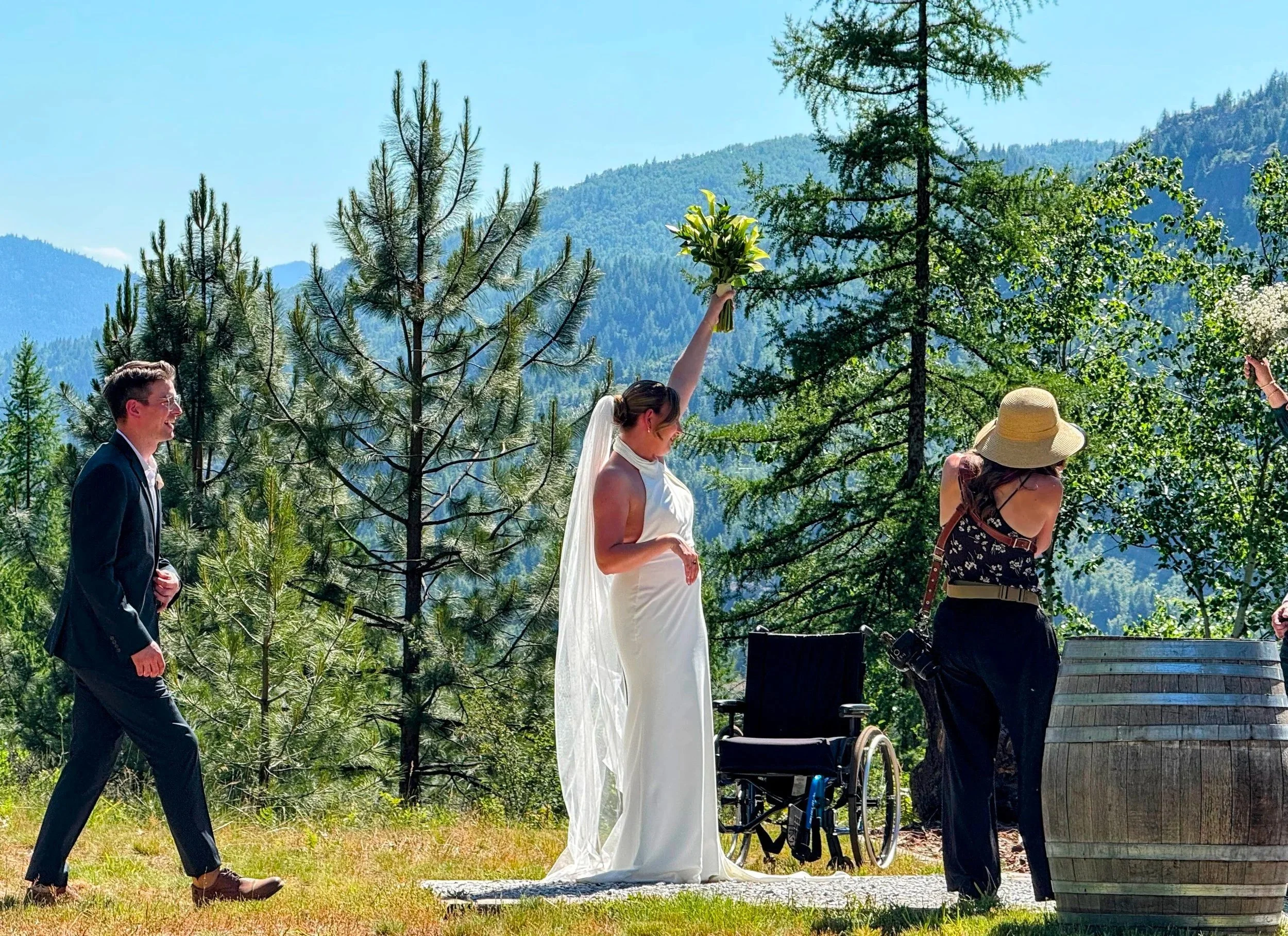 A wedding ceremony taking place outdoors in a forested mountain setting with trees and hills in the background. The bride in a white dress is holding a bouquet high, while a person with a hat stands near a barrel, and a wheelchair is nearby. A man in