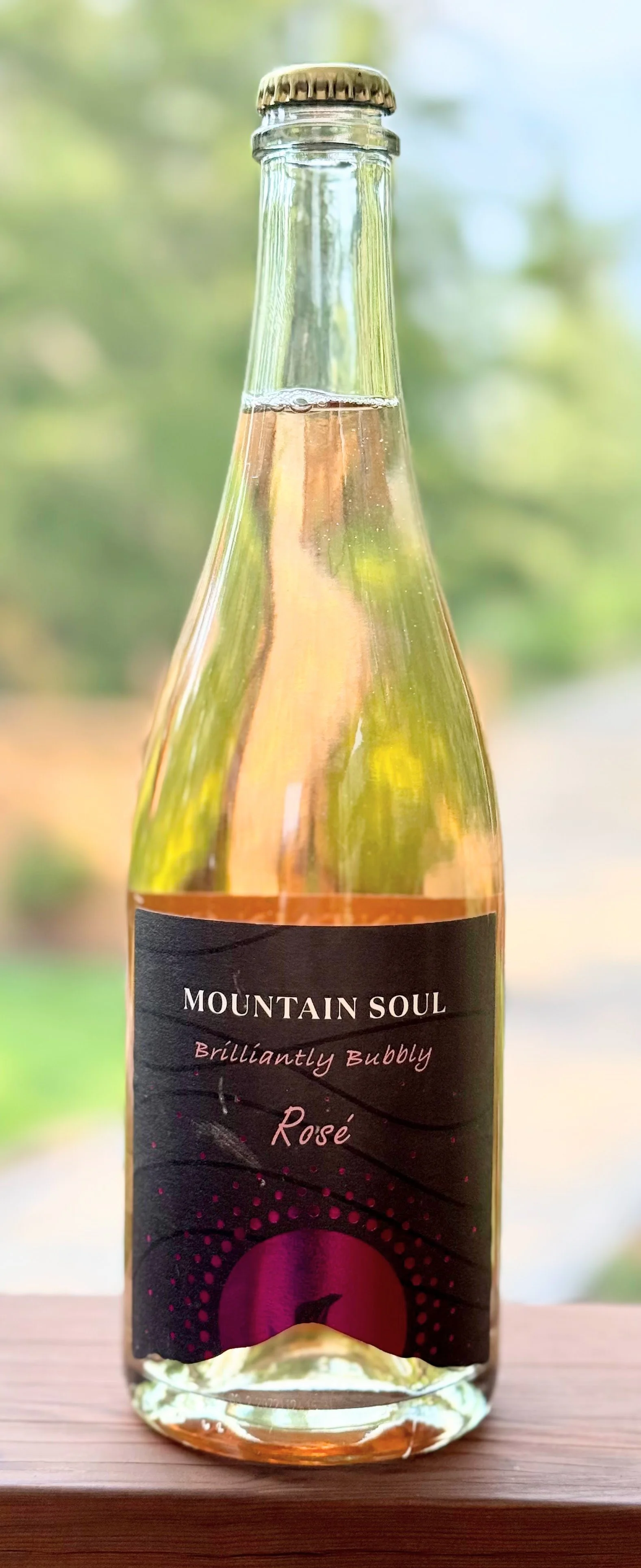 A glass bottle of Mountain Soul Brillanty Bubbly Rosé sparkling wine on a wooden surface, with a blurred outdoor background.