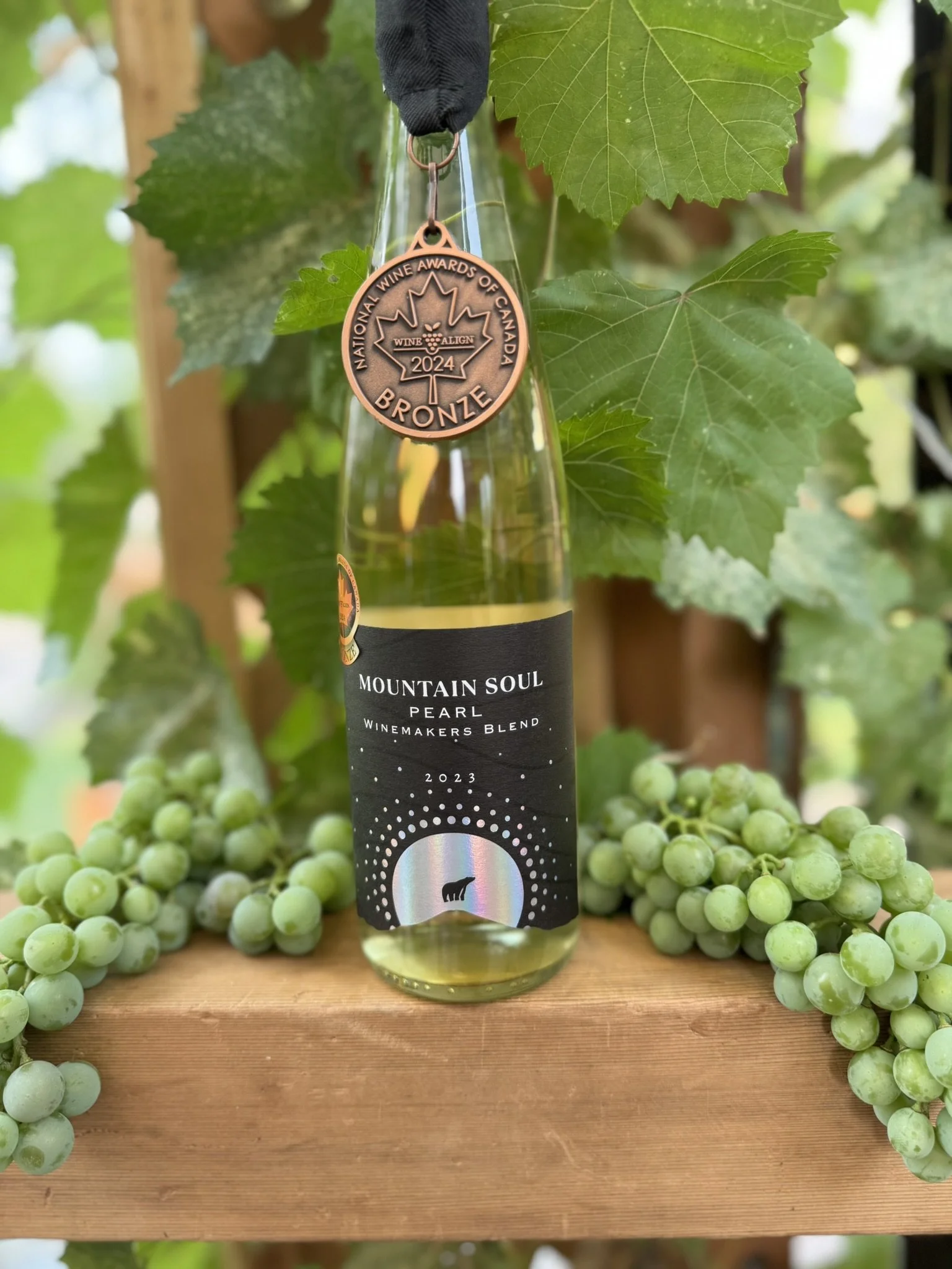 A glass bottle of Mountain Soul Pearl winemaker's blend wine from 2023 is displayed on a wooden surface surrounded by green grape clusters and leaves, with a bronze medal hanging from a black ribbon that reads 'National Wine Awards of Canada Bronze 2024'.
