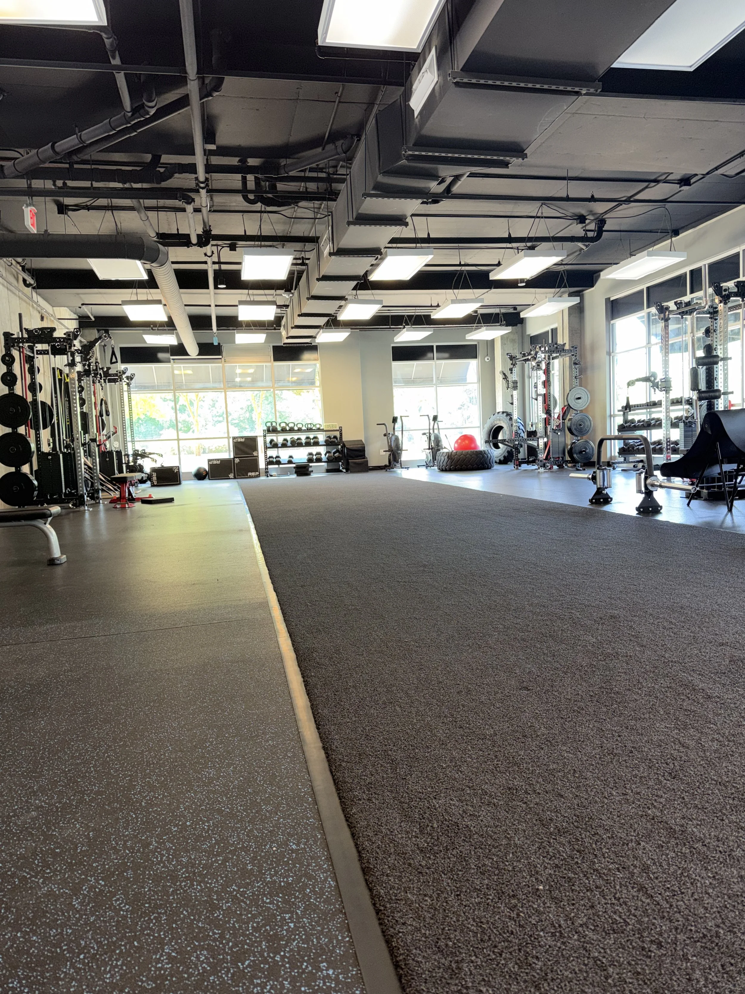 Empty gym with weight racks, kettlebells, a large tire, and cardio equipment near large windows, with a combination of rubber and carpet flooring.