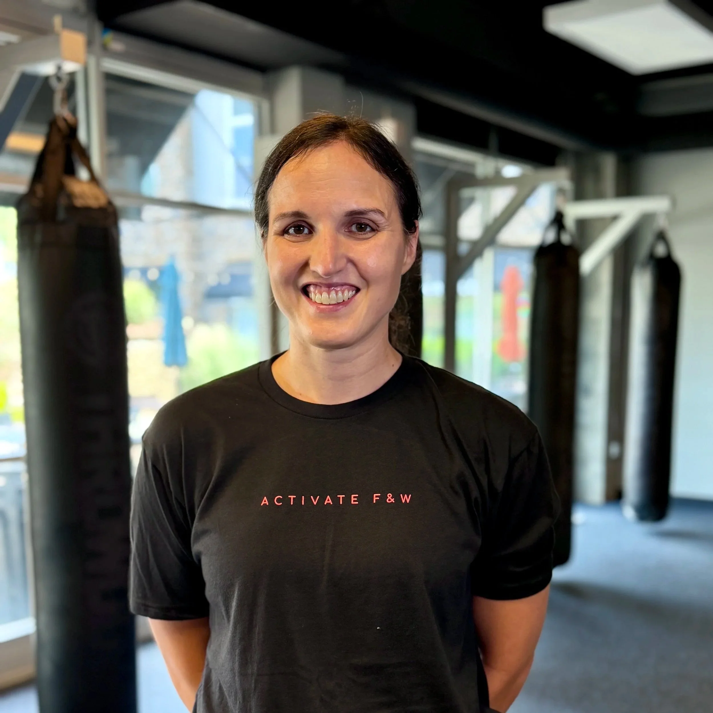 Close-up of a woman smiling at the gym, wearing a black workout shirt with red text, in a gym with punching bags in the background.
