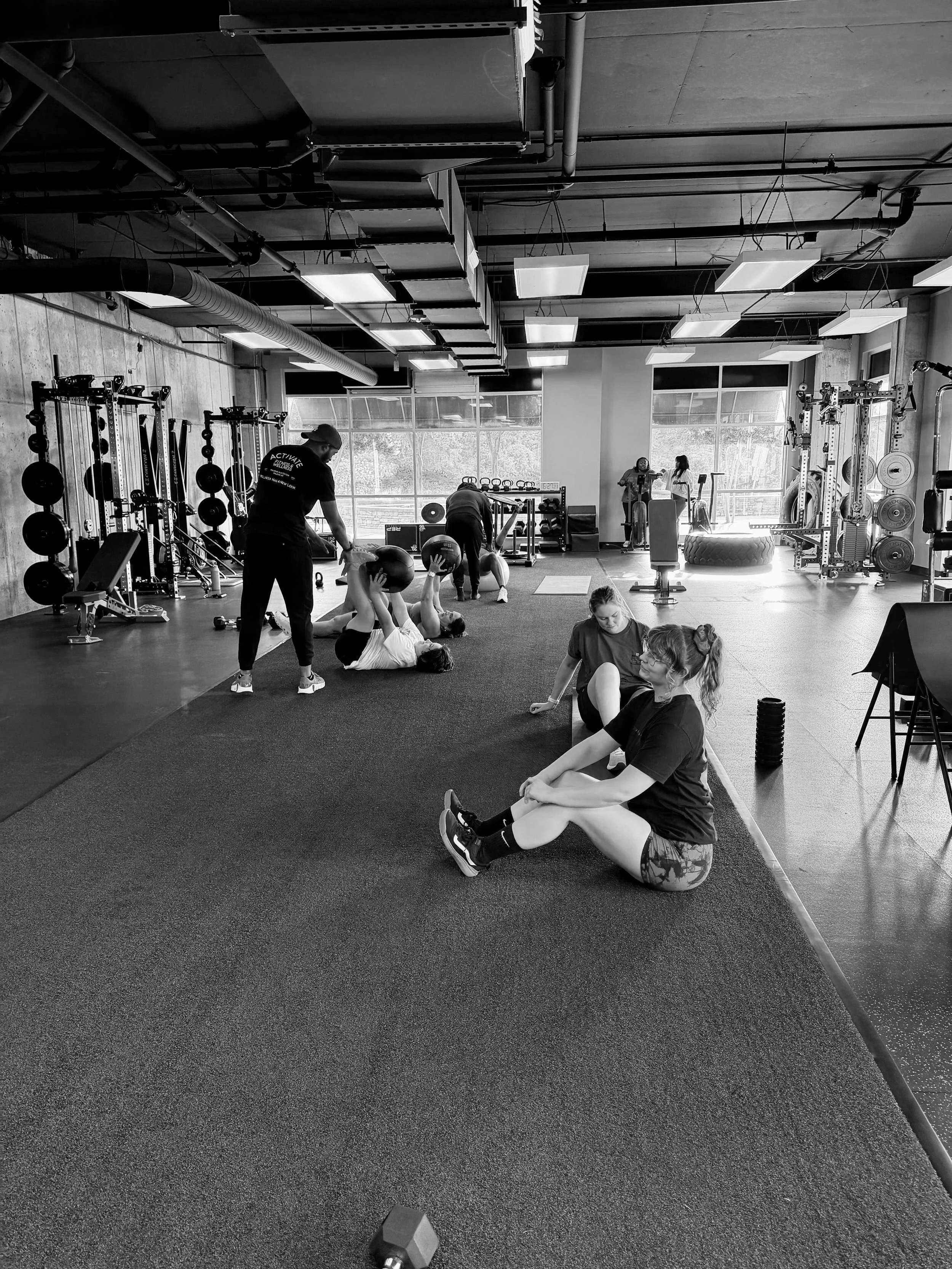 People exercising in a gym, some lifting weights, one person sitting on the floor stretching, and others standing or using equipment, with large windows in the background.