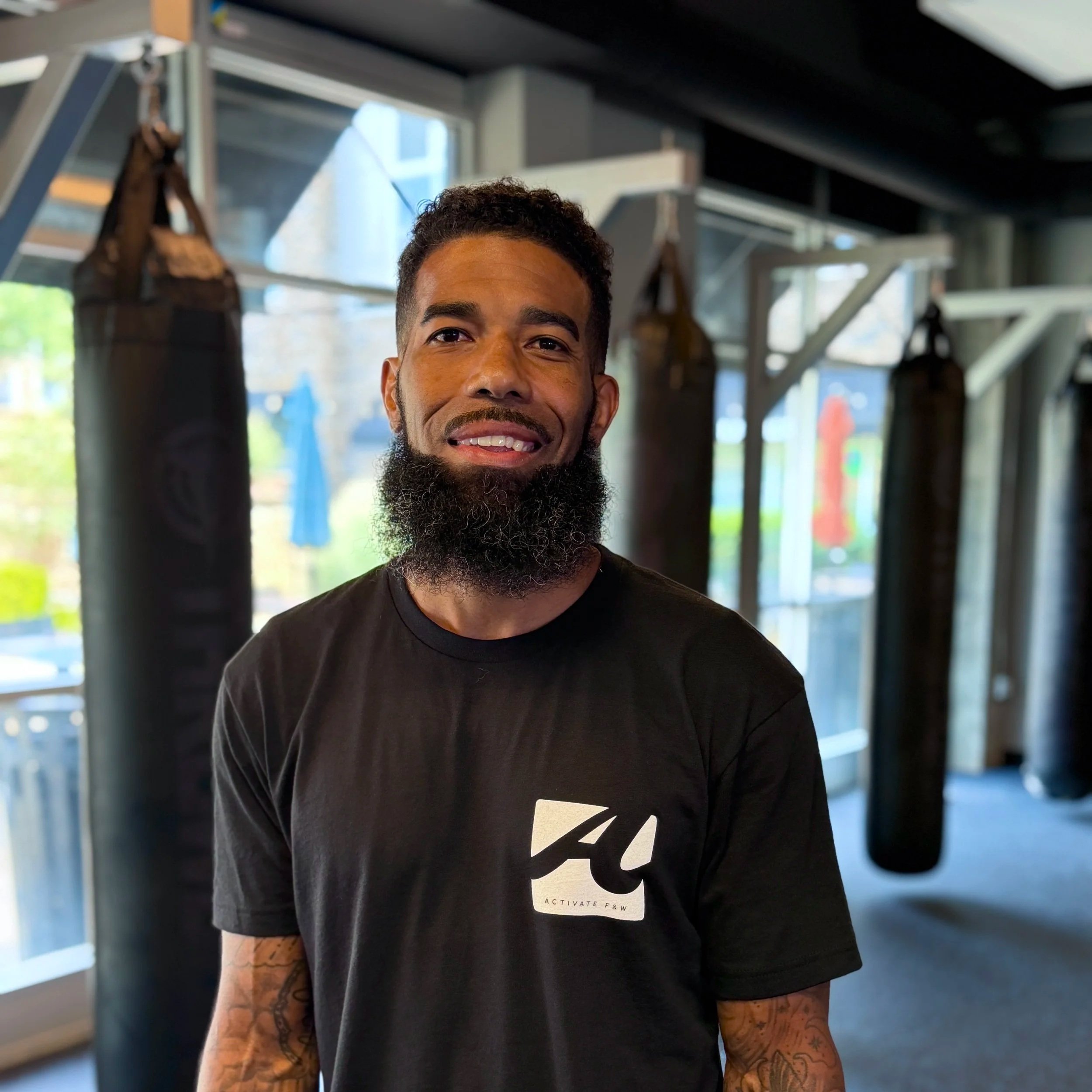 Close-up of a man with short curly hair and a beard, smiling, wearing a black T-shirt with text and graphic, lifting his arms in a gym with punching bags in the background.