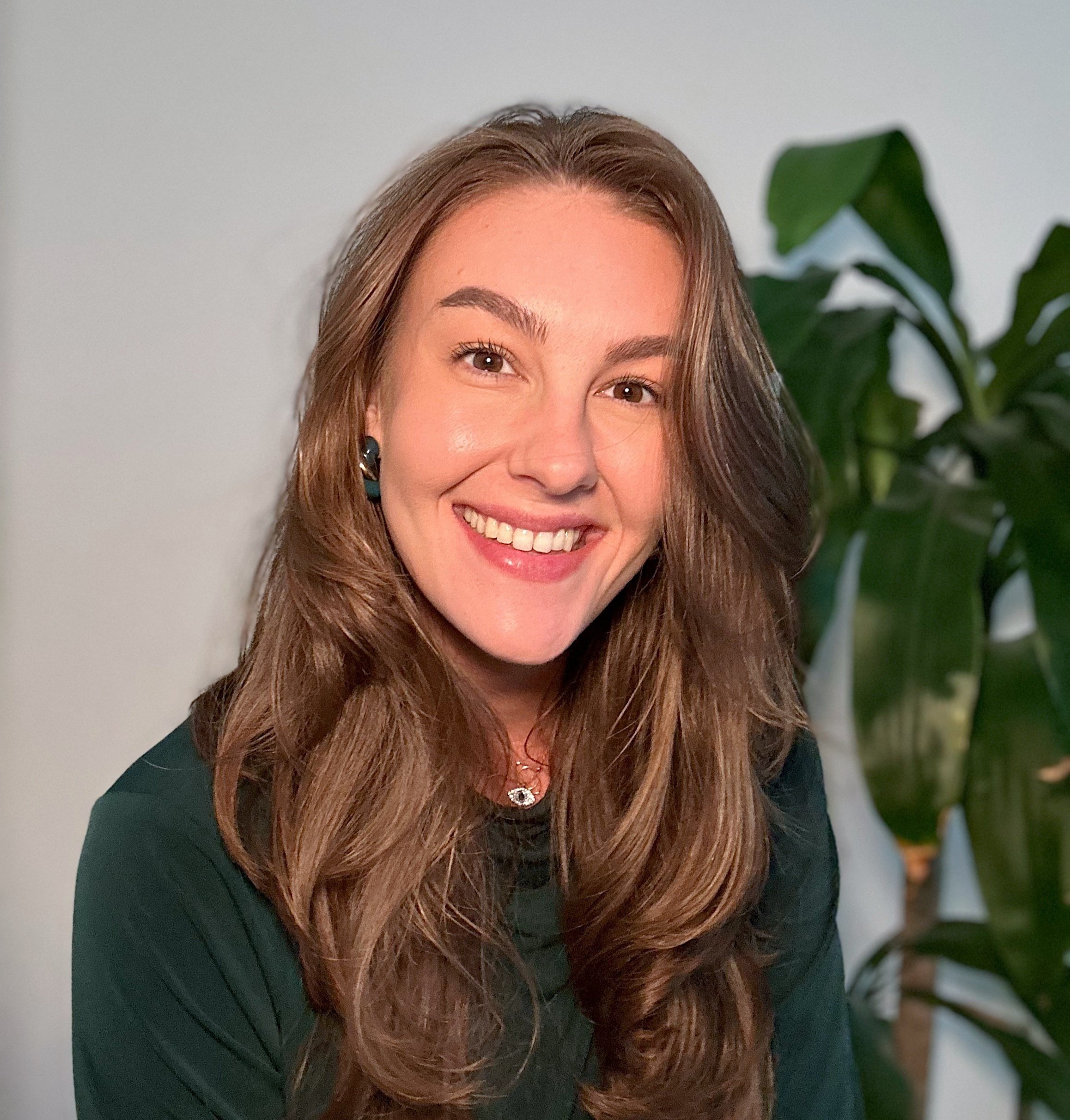 An aspiring writer and avid reader who identifies as a woman with long brown hair wearing a dark green top, standing in front of a white wall and a green plant.