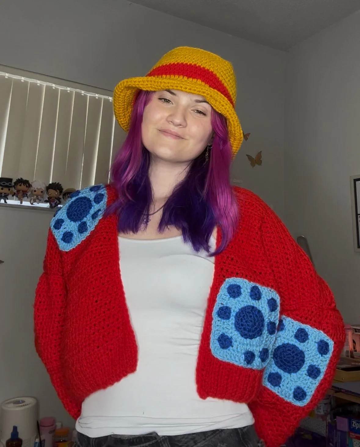 Young woman with purple and pink hair wearing a yellow and red crochet hat and a red crochet cardigan with blue patches, hat is represent Straw Hat Luffy's hat from One Piece. The cardigan represents the Wano outfit Luffy from One Piece wore.