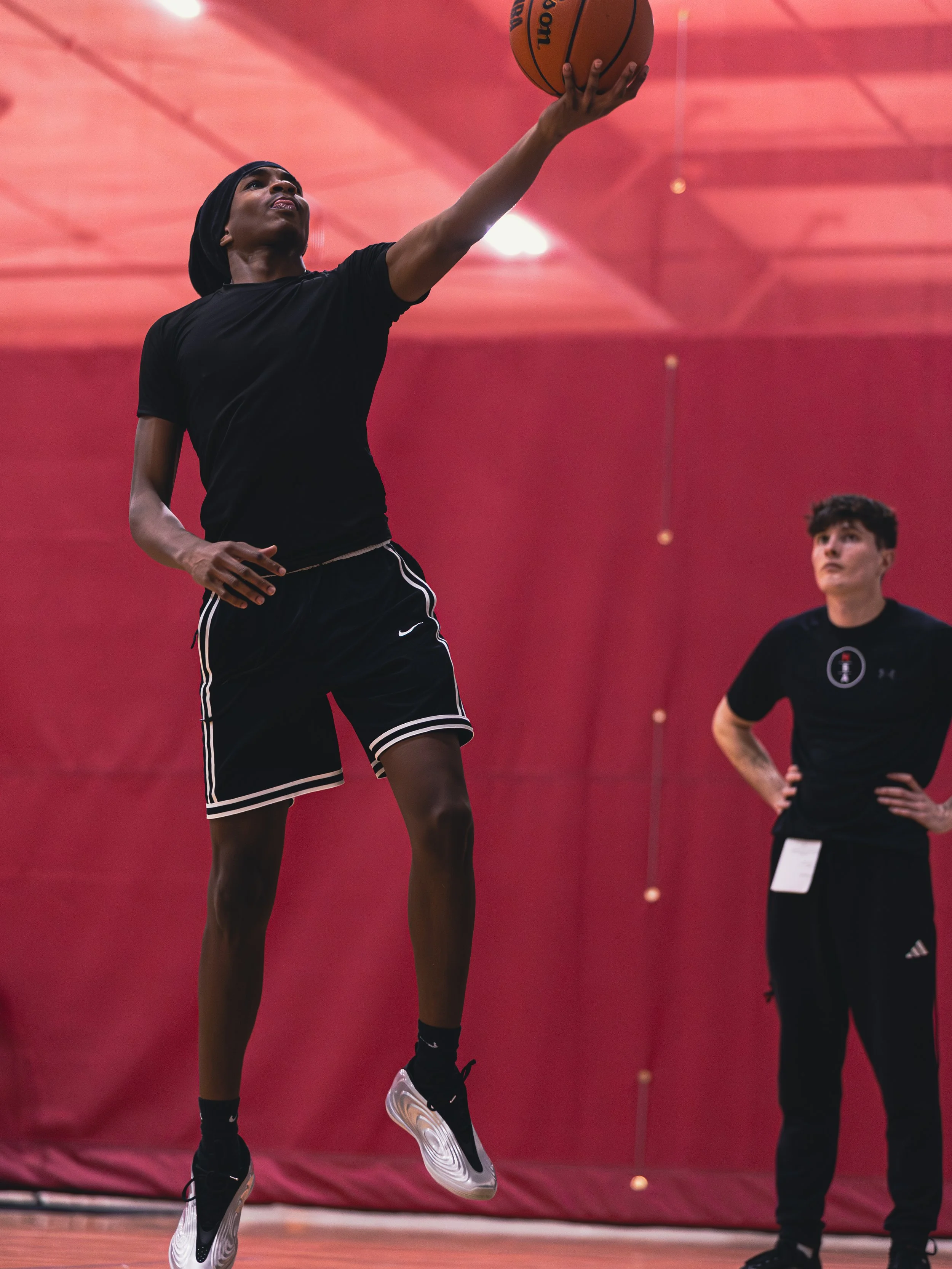 A boy in a black t-shirt and black shorts shooting a basketball on an indoor gym court with a private trainer standing and evaluating nearby