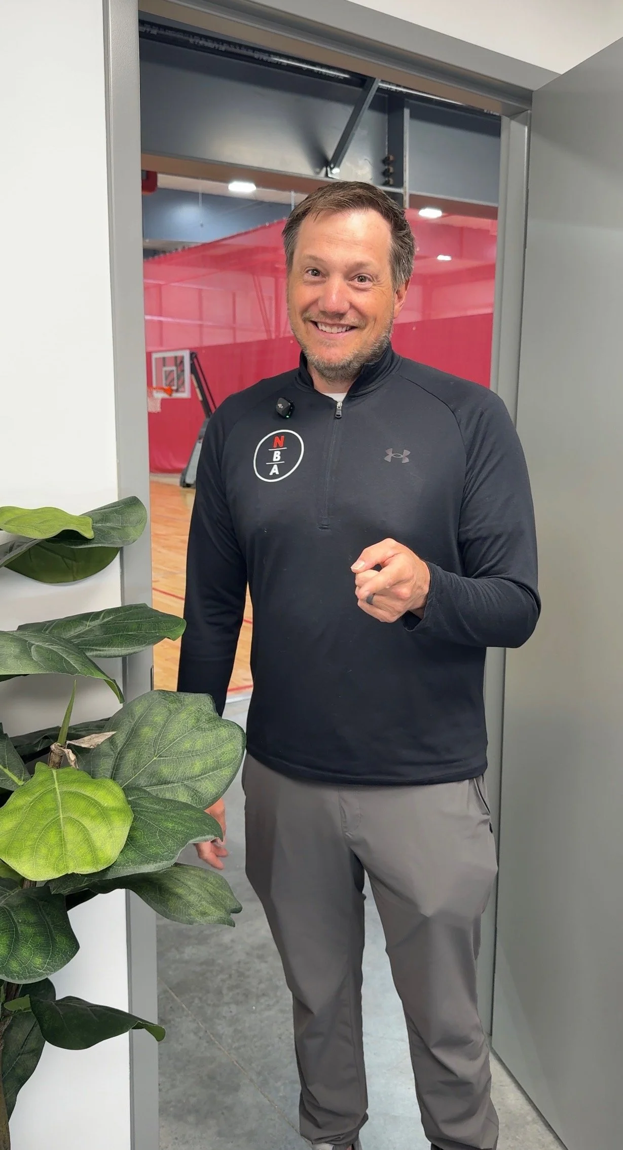 A man in a black jacket holding a Wilson NBA basketball, standing in front of a wall with the Nebraska Basketball Academy logo.
