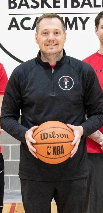 A man in a black jacket holding a Wilson NBA basketball, standing in front of a wall with the Nebraska Basketball Academy logo.