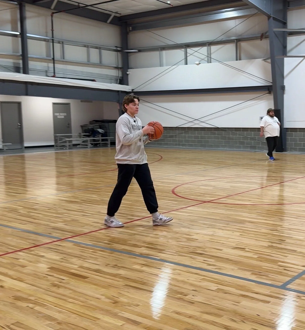 Woman standing in a basketball gym holding a Wilson basketball, wearing a red Nebraska Basketball Academy t-shirt and black shorts, with a Nebraska Basketball Academy logo in the background.