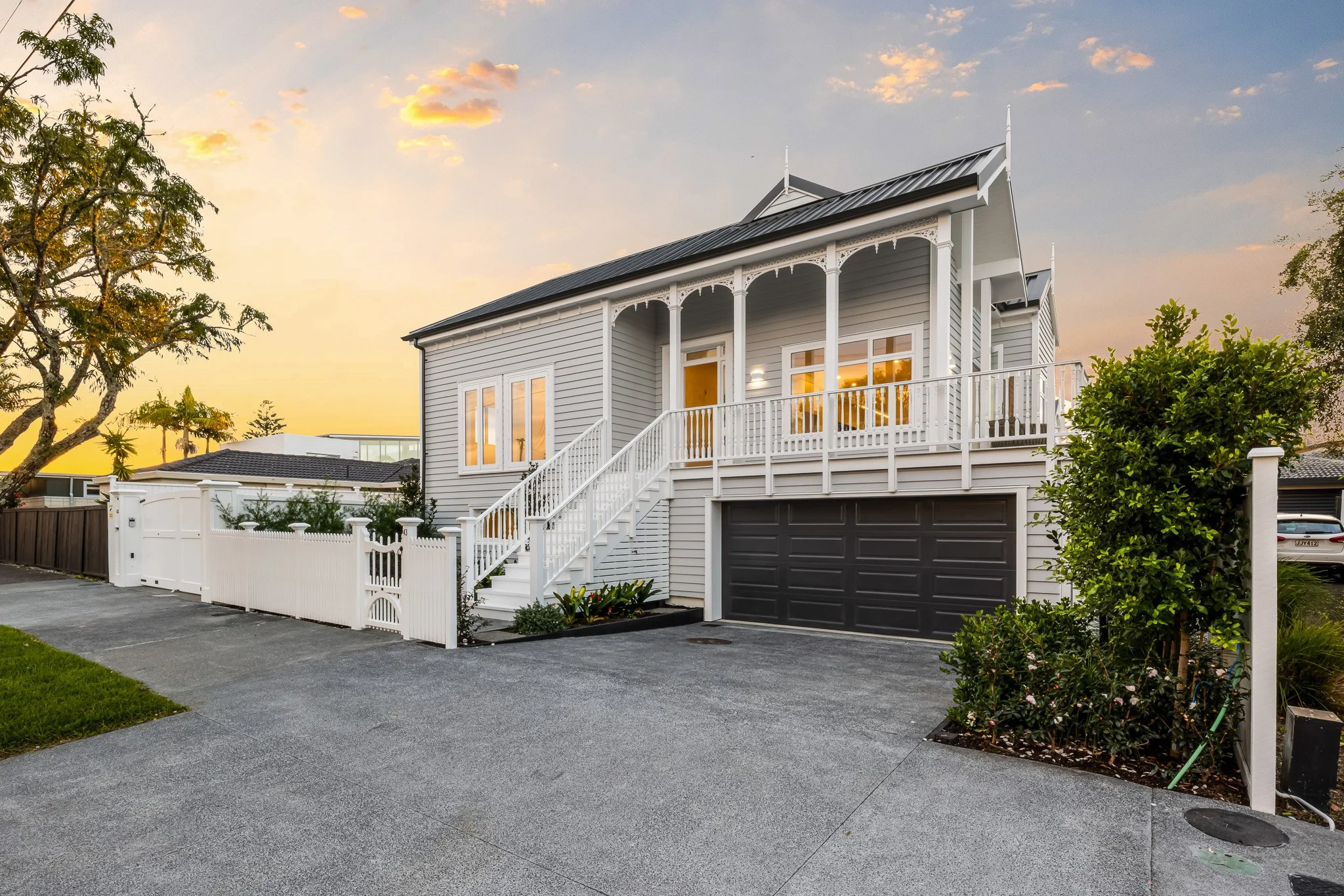 Modern two-story house with white picket fence and garage, surrounded by garden, against sunset sky.