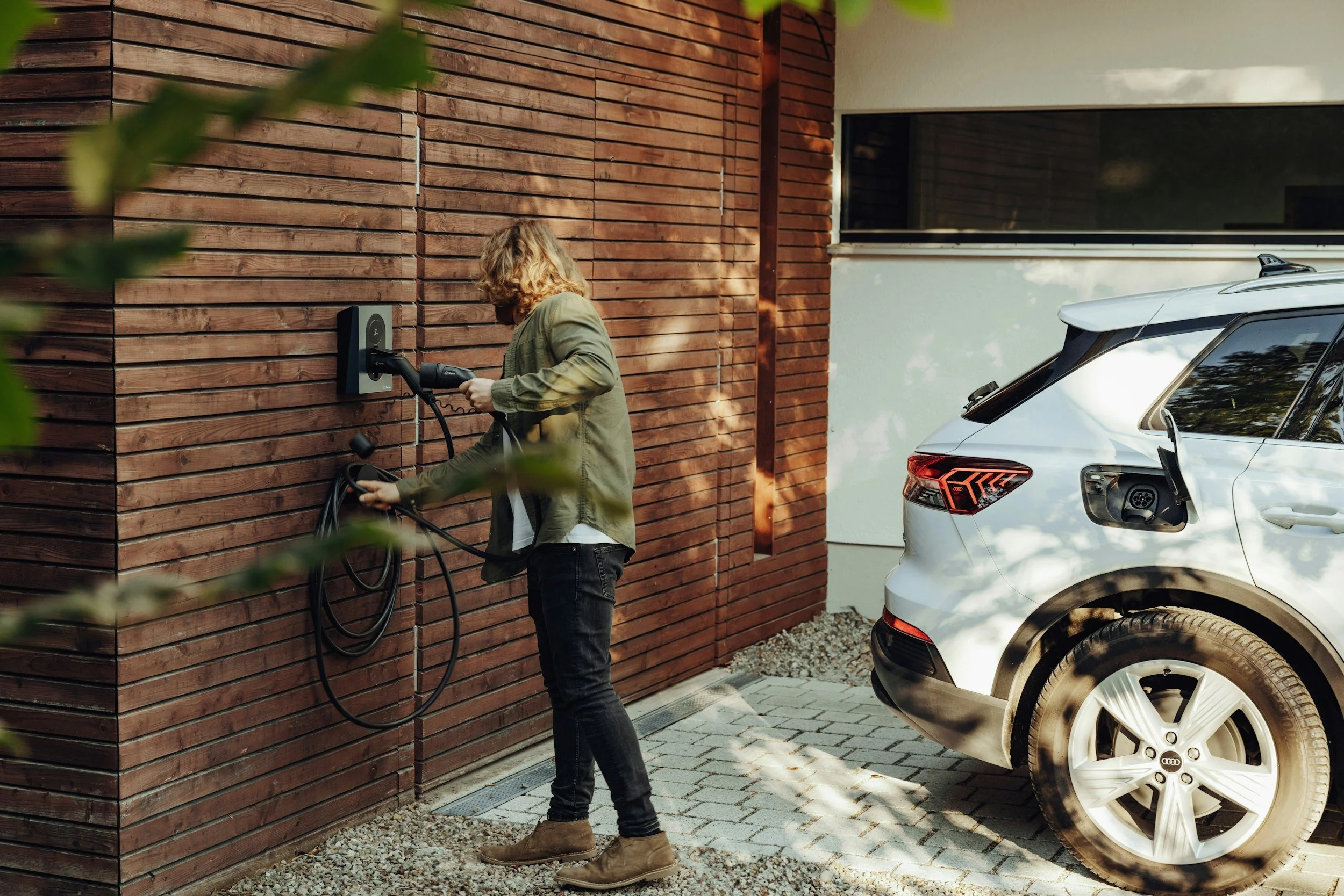 A person charging an electric car at a house driveway with a wooden wall and a white vehicle nearby.