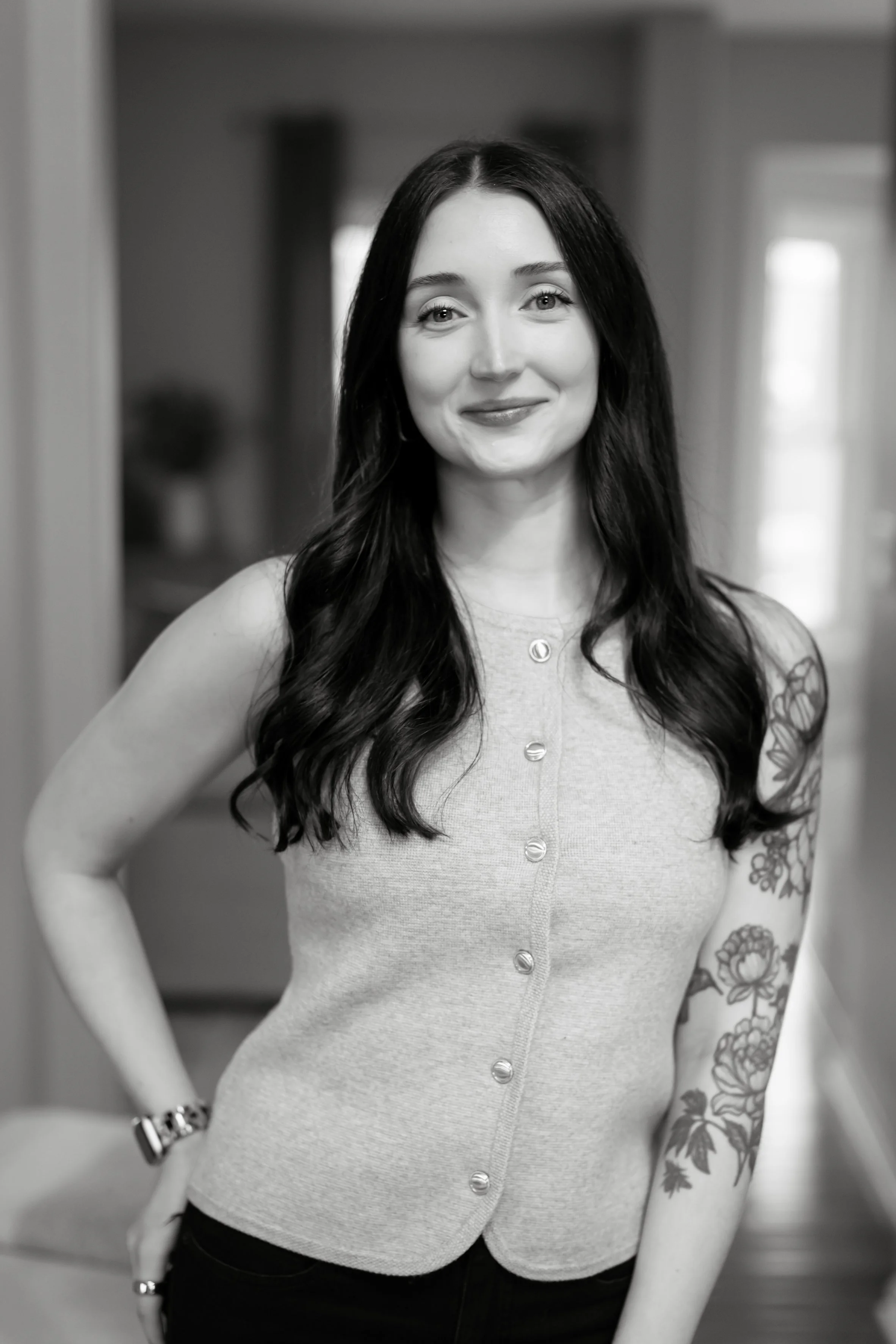 Black and white photo of a woman with long dark hair, smiling slightly, wearing a sleeveless button-up top and a watch on her left wrist, standing indoors with a blurred background.