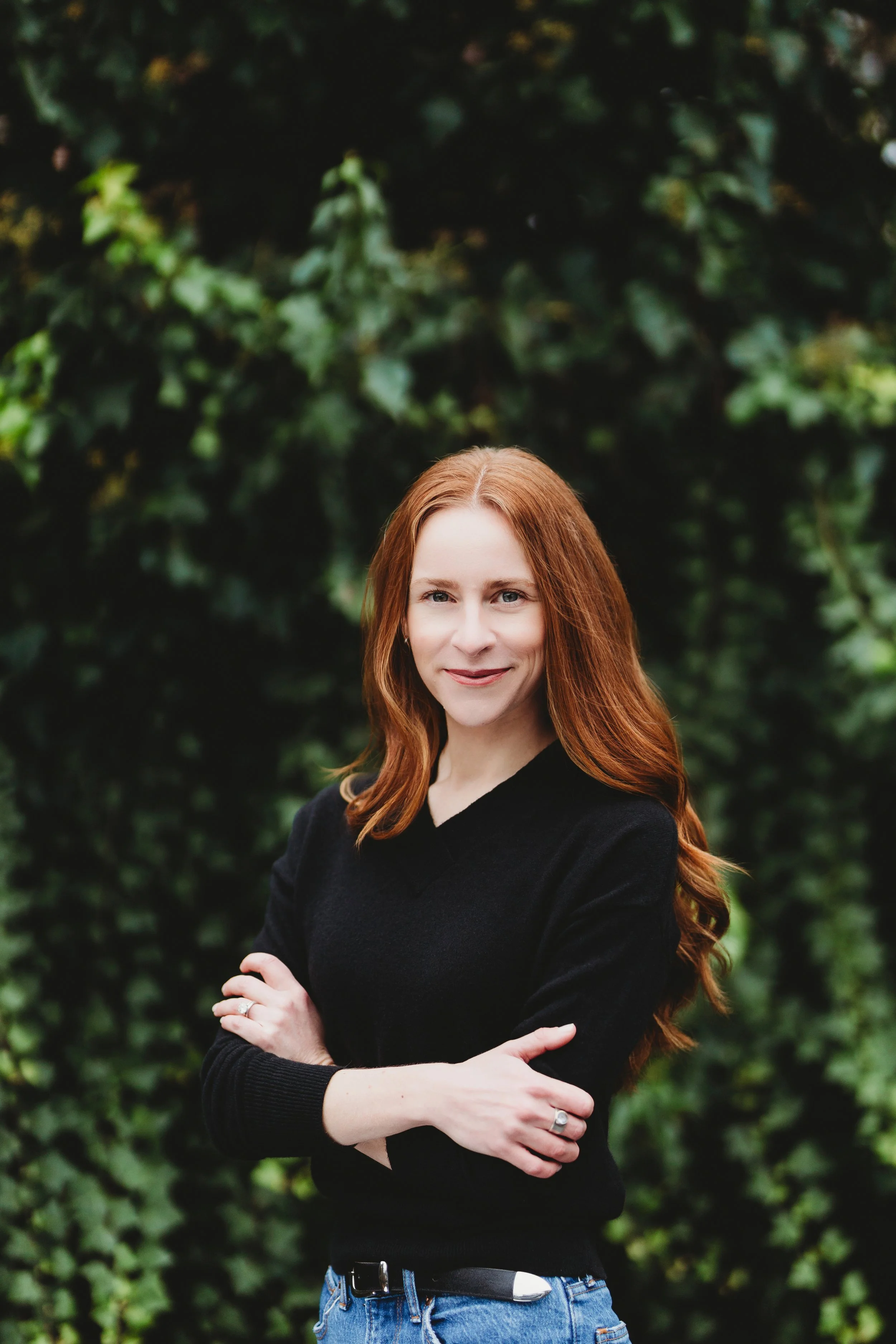 A smiling woman with red hair, blue eyes, and fair skin, outdoors with blurred green foliage in the background.
