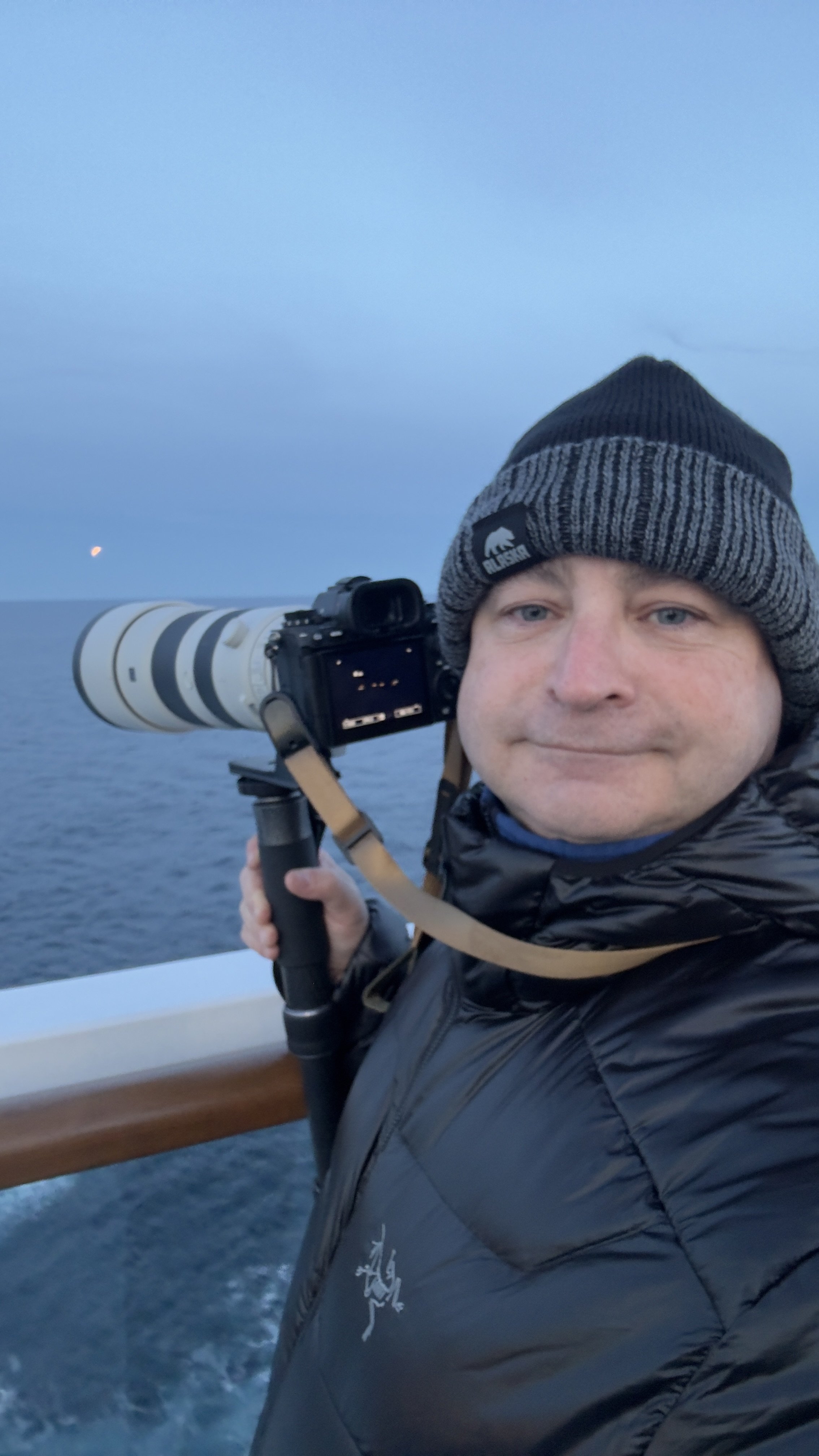 A man in a black jacket and gray knit beanie holding a large camera with a long telephoto lens outdoors on a boat, with water and a sky in the background, and a small crescent moon visible in the sky.