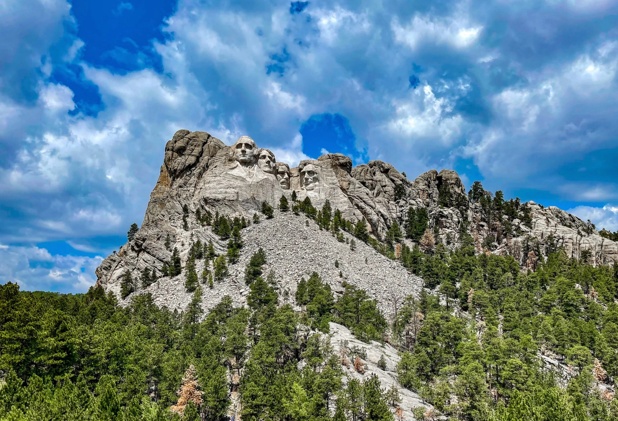 Mount Rushmore above a forested slope beneath large white clouds