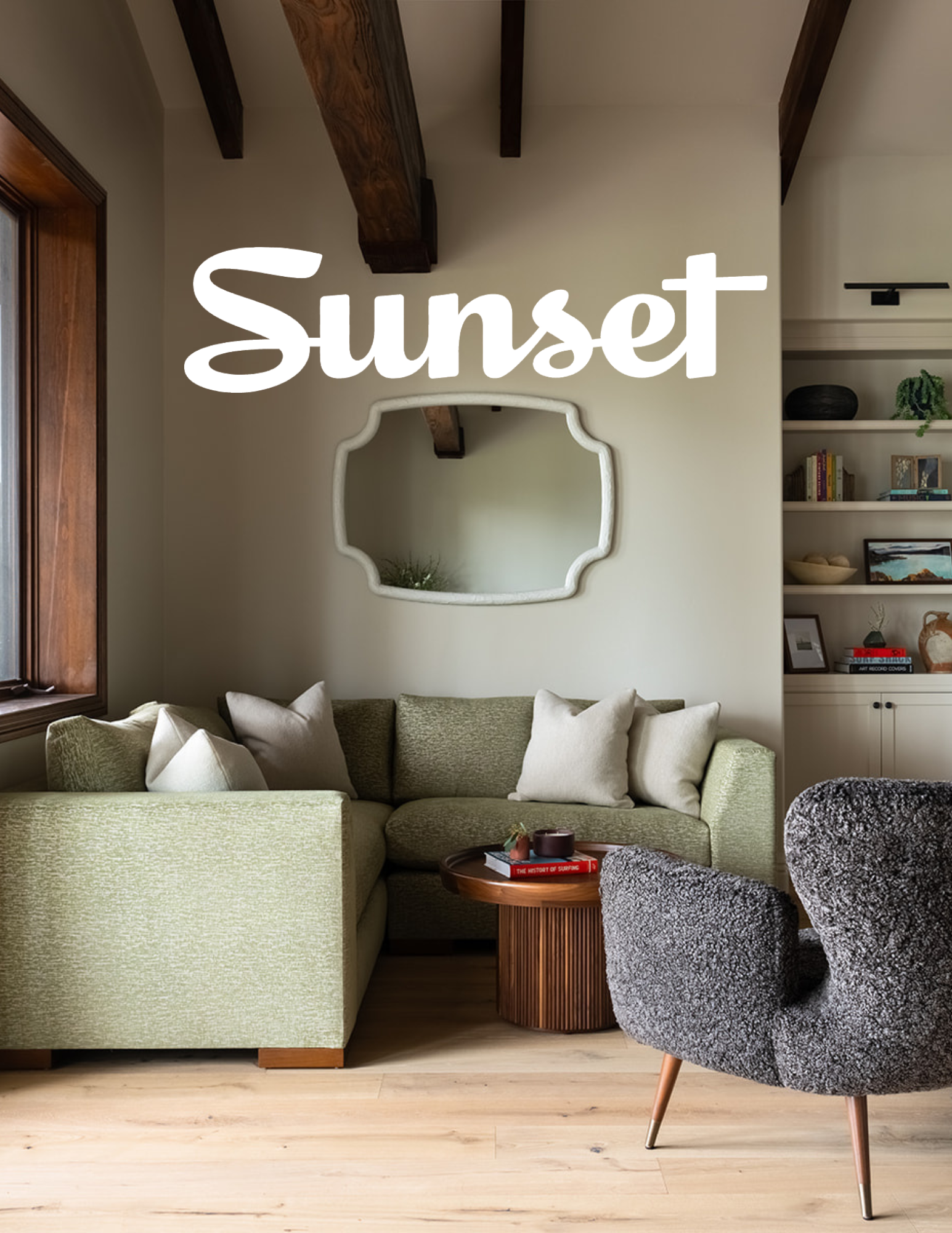 Living room with green sectional sofa and gray armchair, wooden side table, white wall mirror, and built-in bookshelf, illuminated by sunset light from a window.