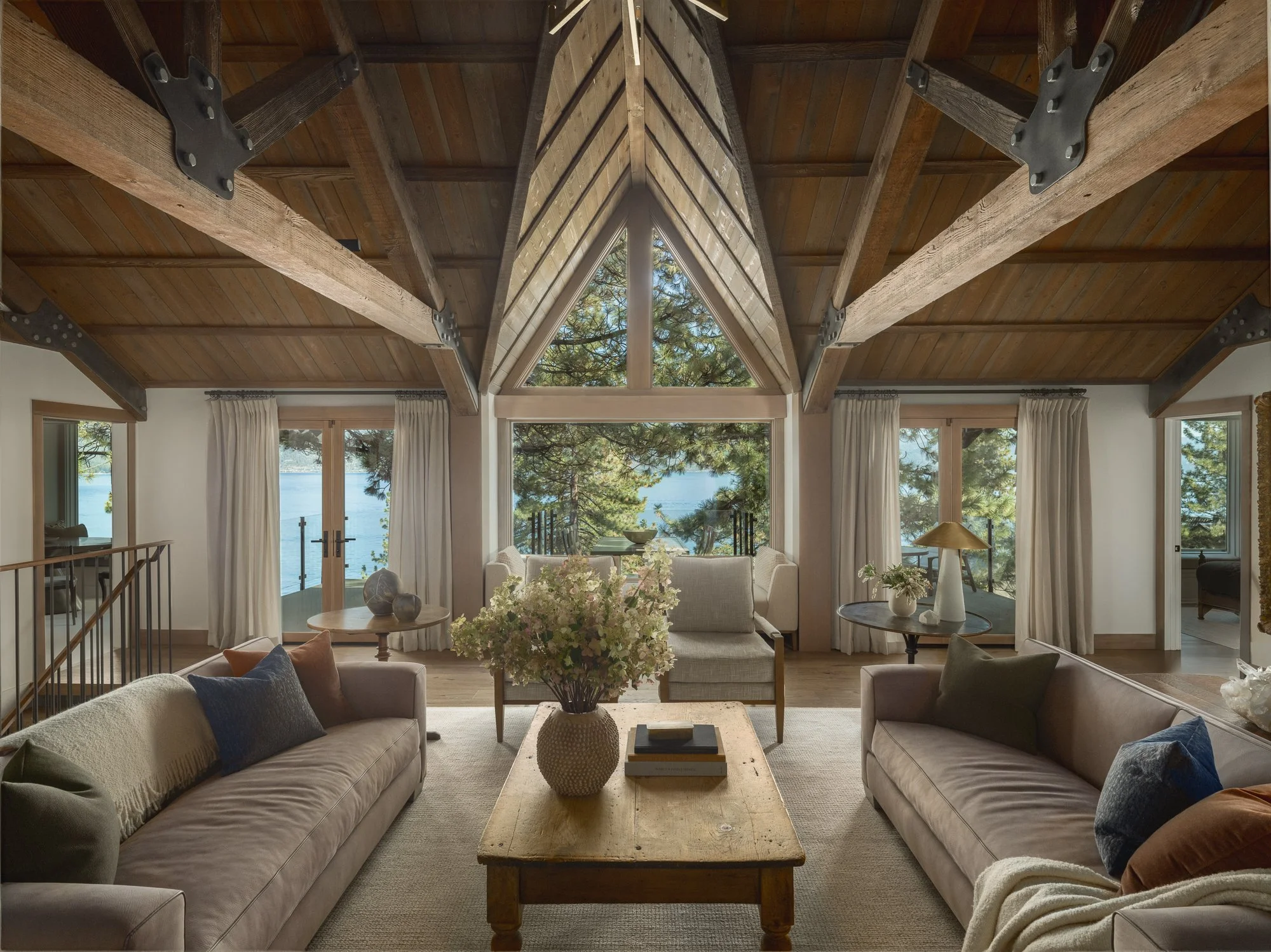 Transitional mountain living room with vaulted wooden ceiling, large central window with views of Lake Tahoe through trees, sofas with textured and earthy colored pillows, vintage wooden coffee table with a vase of flowers, and custom upholstery. Des