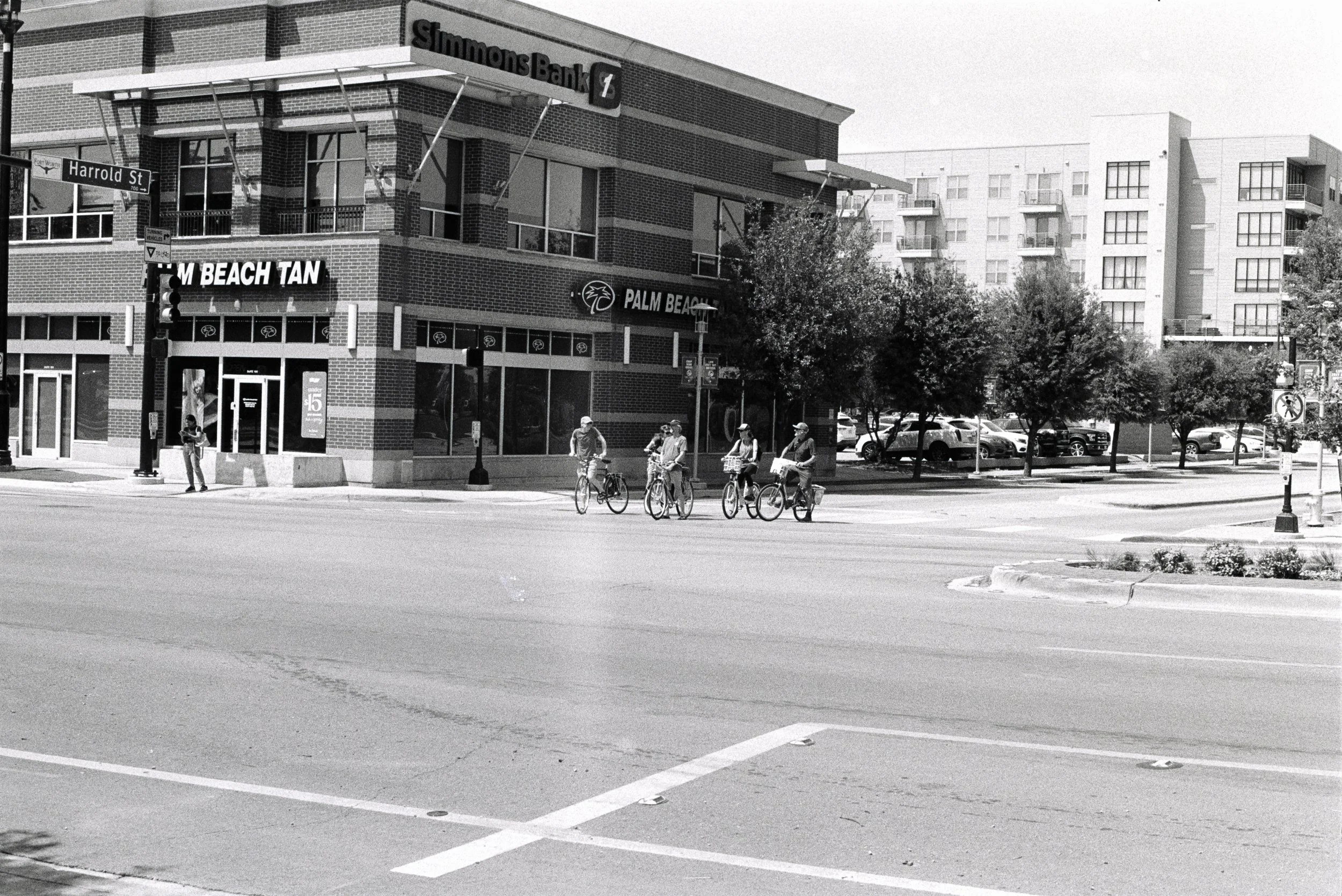 A street corner with a large brick building housing a Simmons Bank and a Palm Beach tan store. Four people are riding bikes at the crosswalk, and one person has stood on the sidewalk with a phone. Several trees line the parking lot with cars parked i