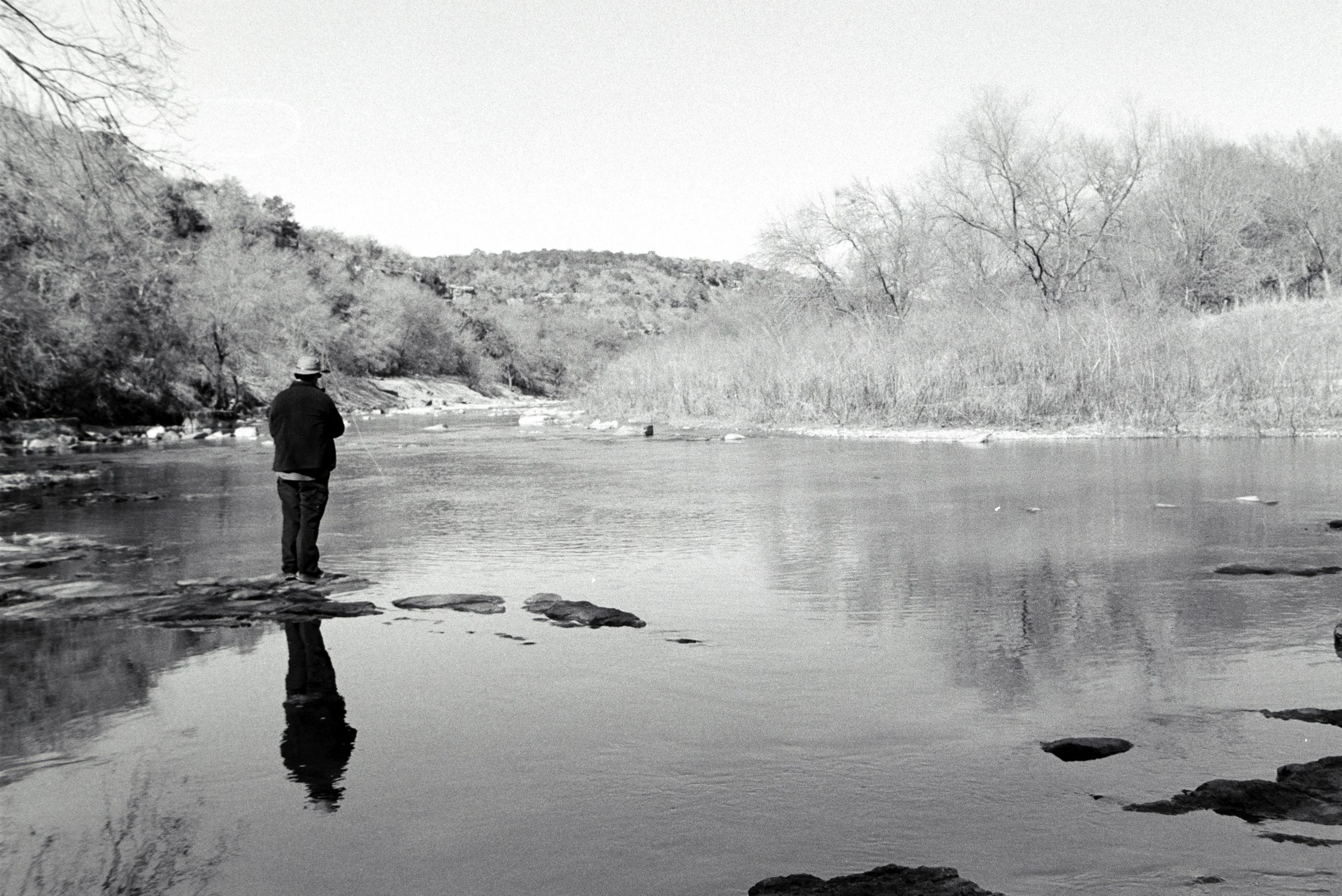 A person in a hat standing and fly fishing in a river with trees and hills in the background, in black and white.
