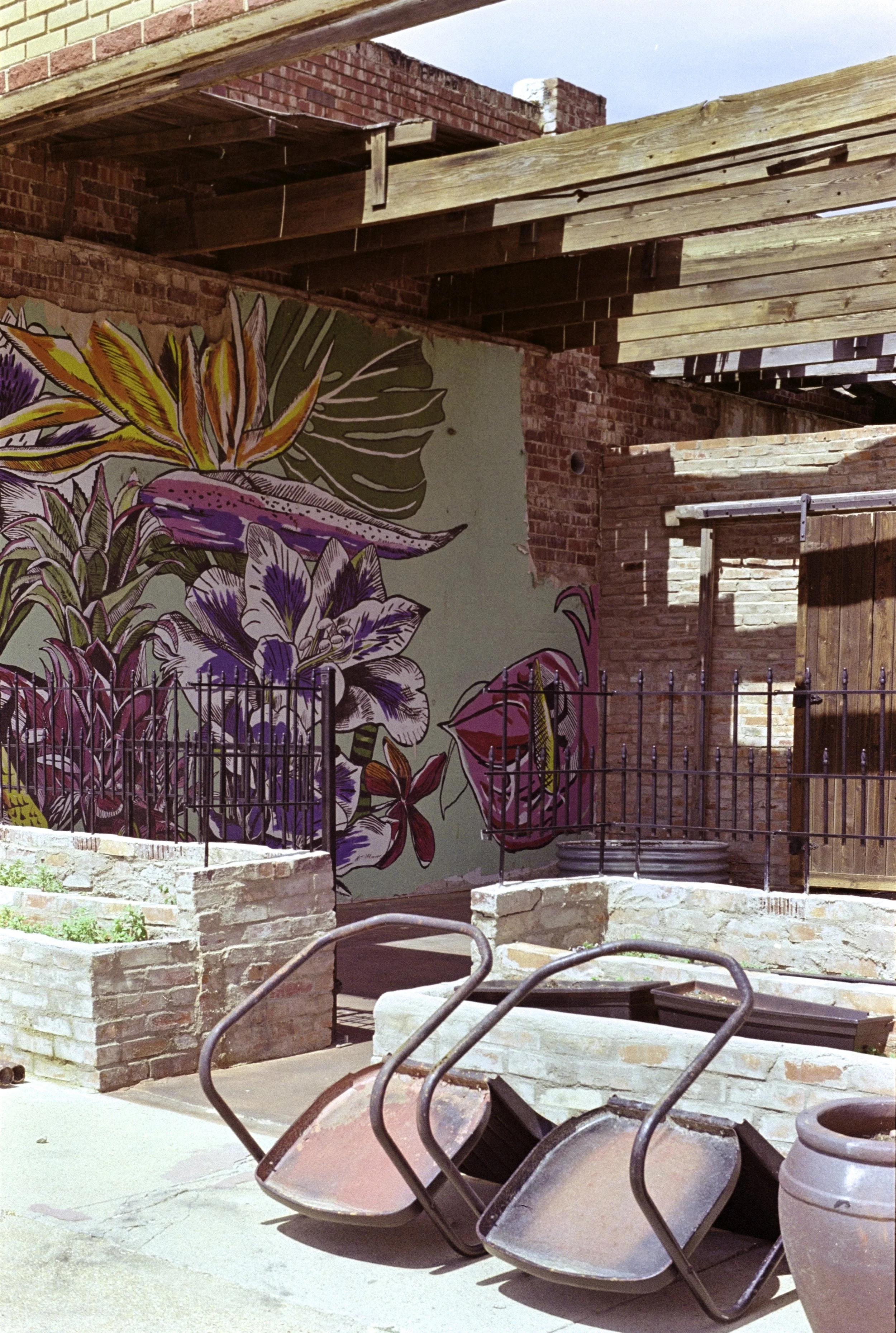 Rusty upside down chairs and large clay pots in front of a mural with colorful flowers and leaves, brick walls, and wooden beams.