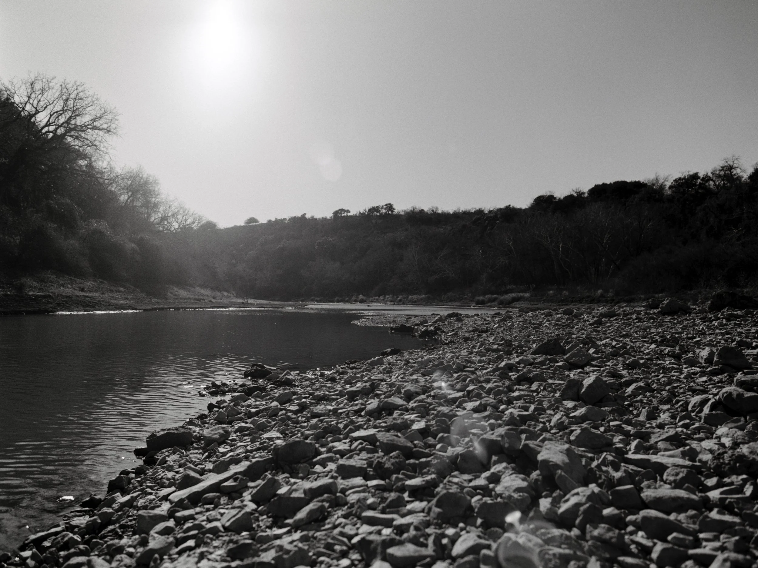 A river flowing through a forested landscape with leafless trees and a bright sky overhead.