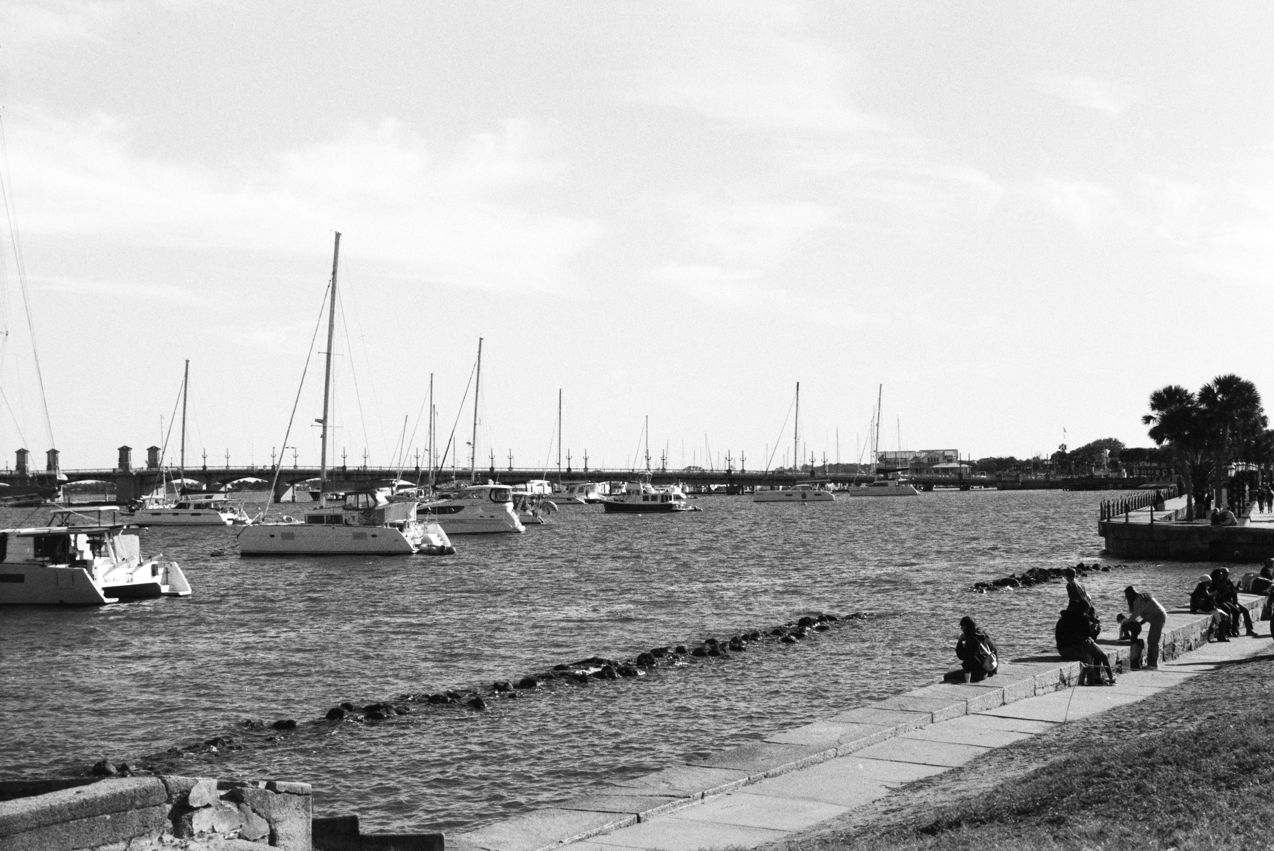 People sitting on a concrete shoreline and walking along a paved path by a marina with sailboats and yachts docked, and a bridge in the background.
