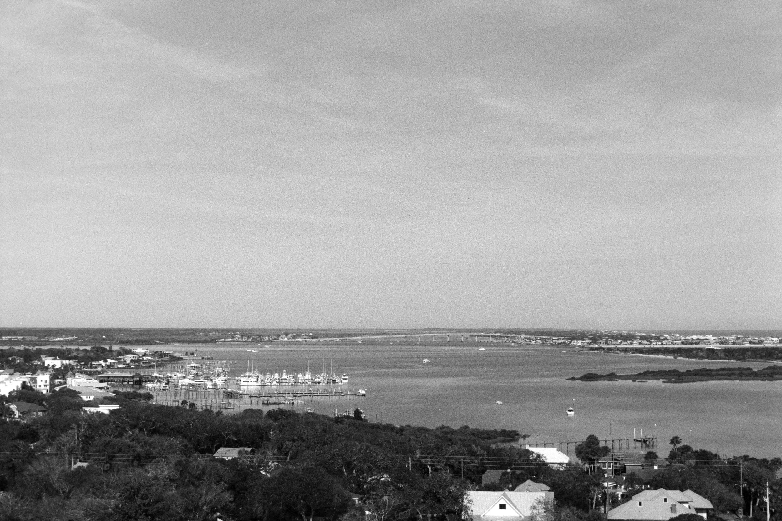A black-and-white photo of a coastal cityscape showing houses, buildings, boats, and a body of water with a bridge in the distance.