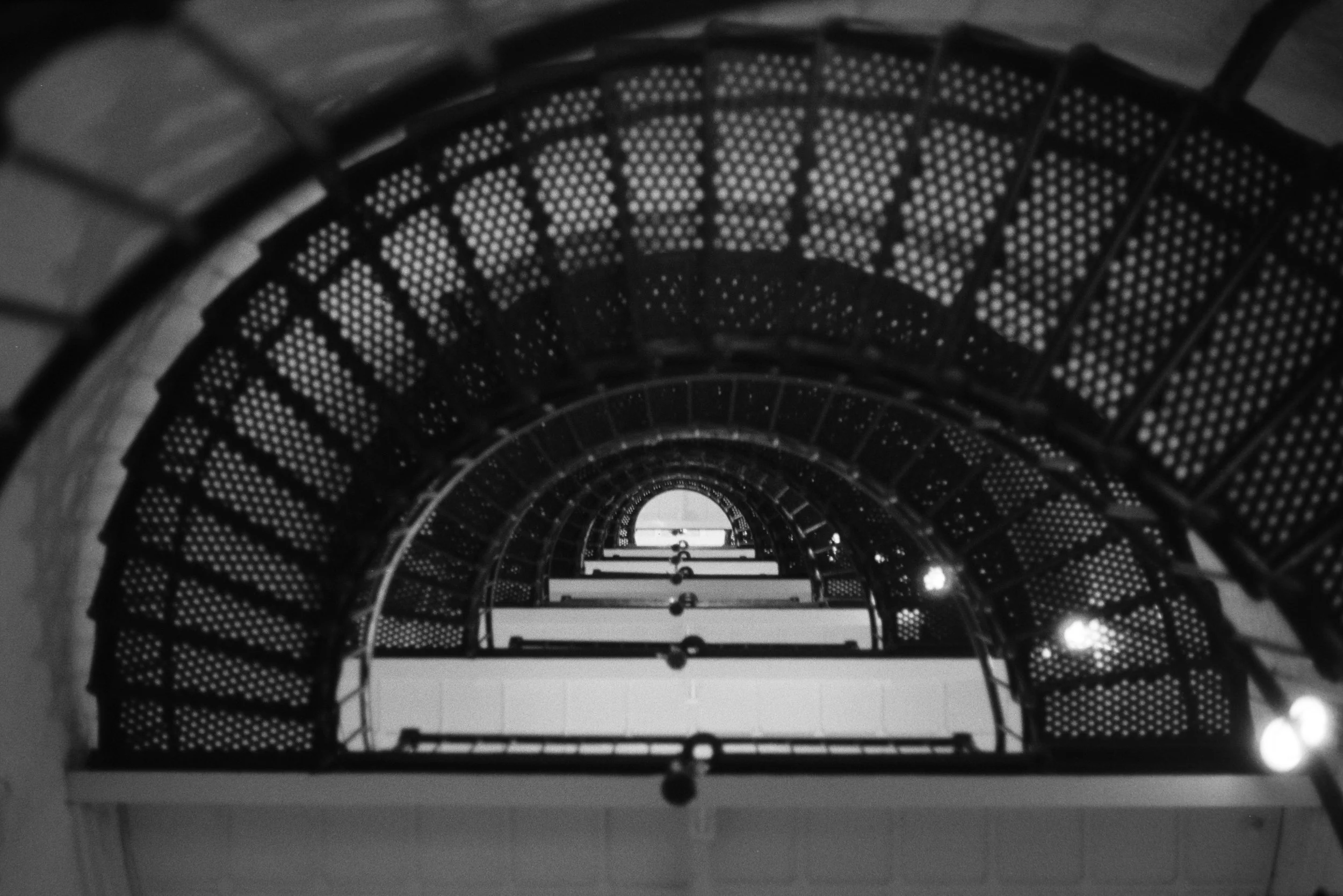 Black and white photo capturing the inside view of a spiral staircase with perforated steps, creating a tunnel-like perspective upward.