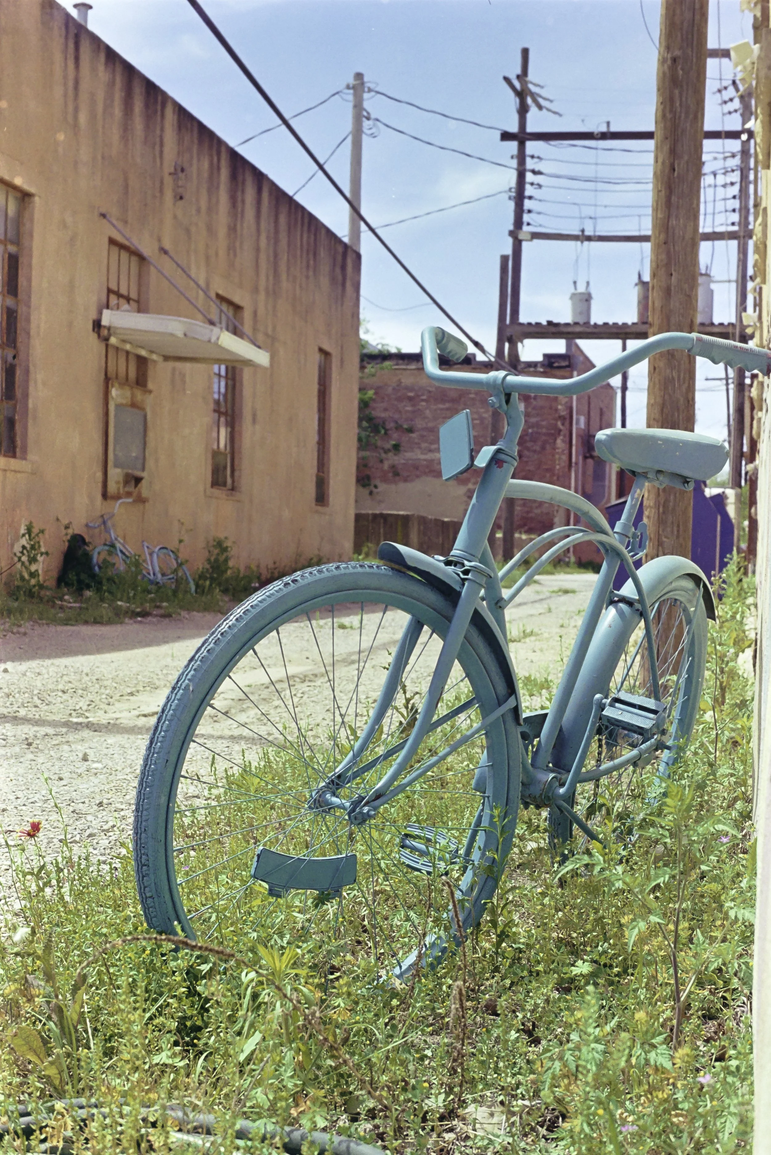 A light blue bicycle parked on a patch of grass and weeds in an alleyway. In the background, there are weathered brick buildings and utility poles with overhead power lines under a blue sky.