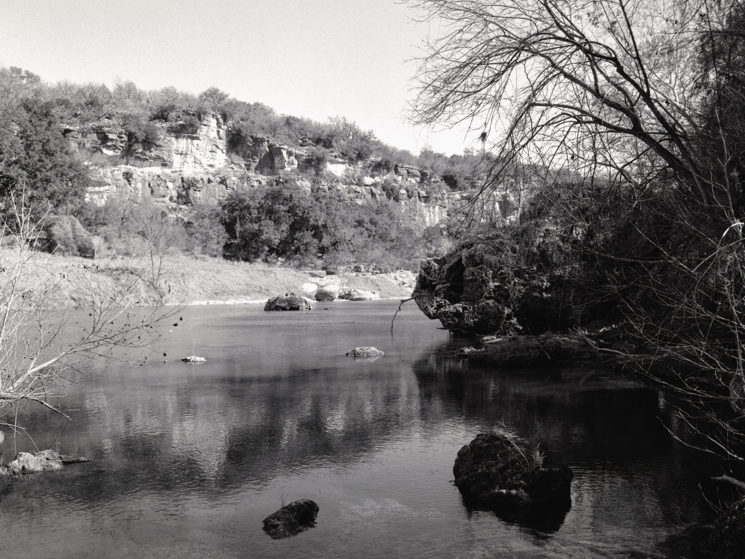 Black and white photograph of a river with rocks, trees on the riverbank, and a rocky hillside in the background.