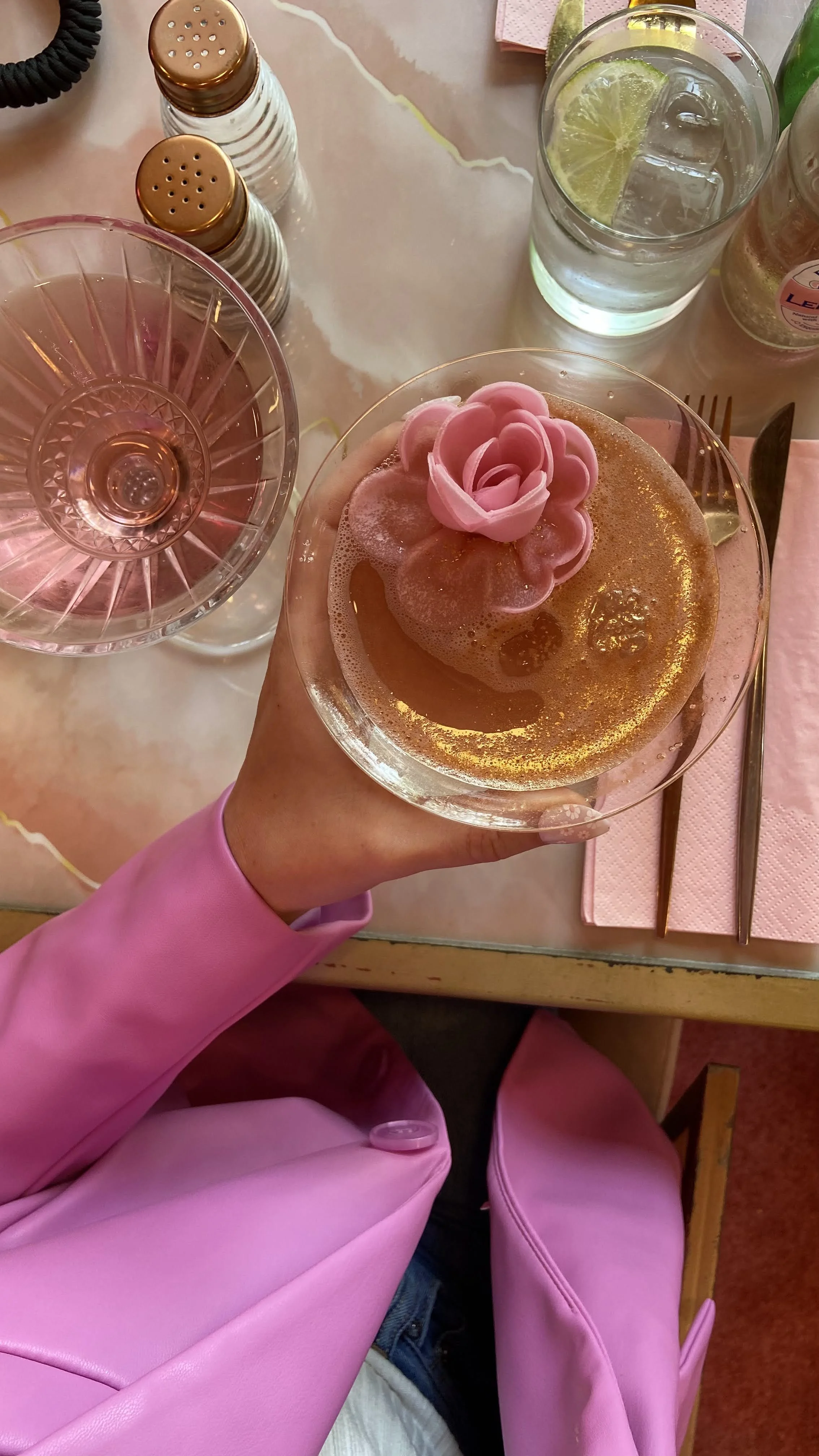 A person holding a cocktail with a pink rose garnish, sitting at a table with a water glass with lime, pink napkin, and salt and pepper shakers. The person is wearing a pink jacket.
