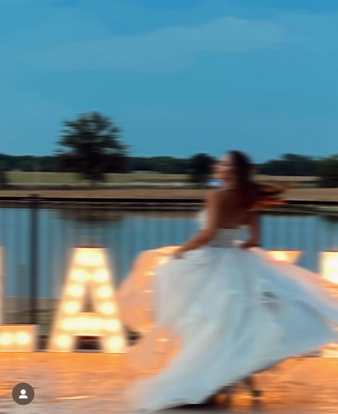 A blurred image of a person in a white dress twirling outdoors near a fence with illuminated letters in the background.
