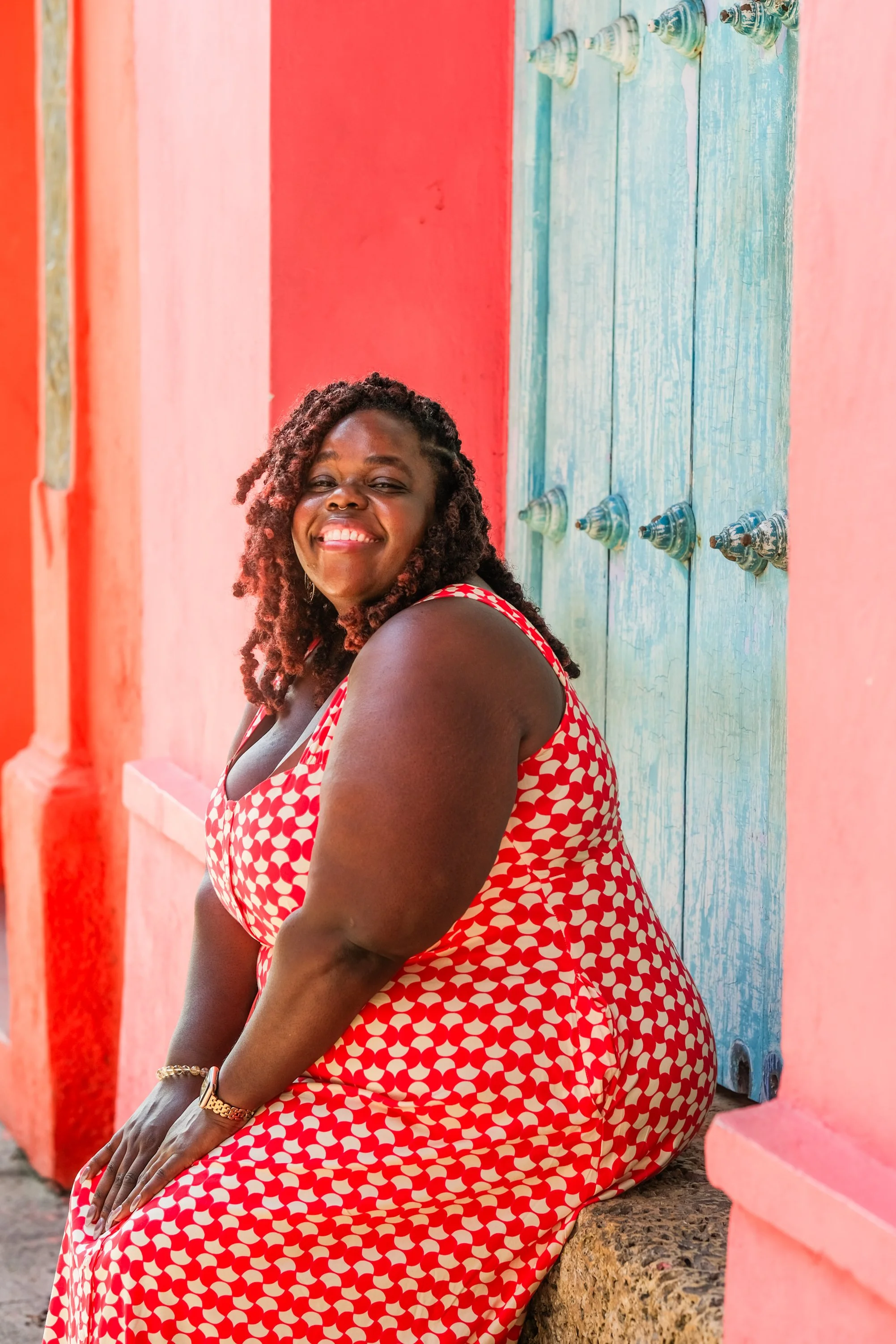 Lauren Hill, a wedding officiant, is seated on a stone ledge in Cartagena, Colombia, surrounded by vibrant colors. She is wearing a red and cream patterned dress and smiling radiantly.  The backdrop features a brought coral wall and a rustic door.