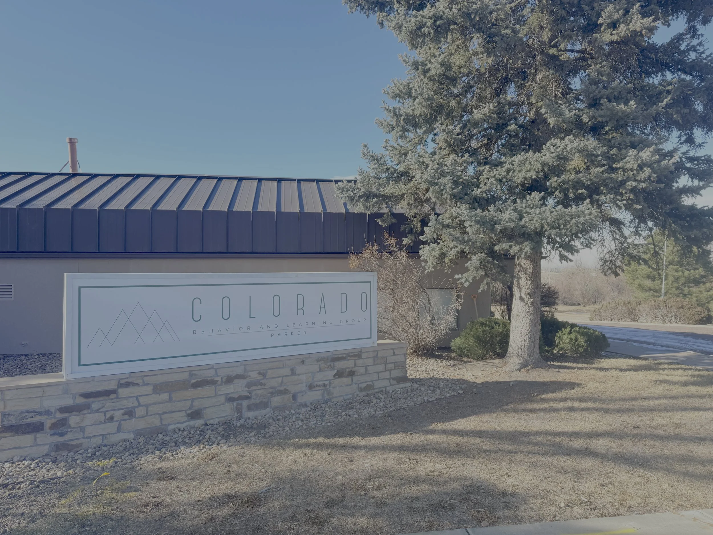 Sign reading 'Colorado Behavior and Learning Group Parker' in front of a building with a black metal roof, pine tree, and bushes.