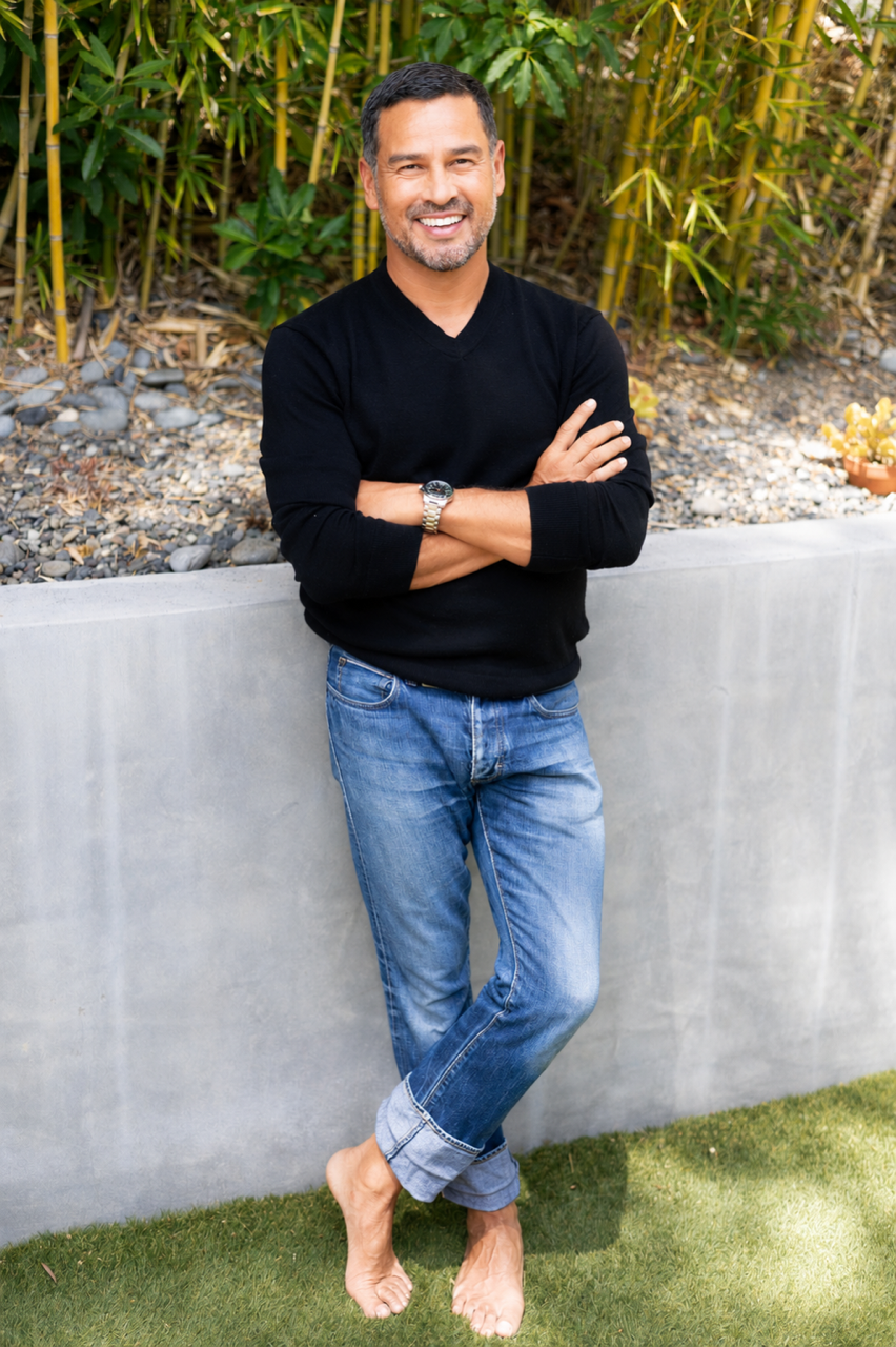 A smiling man with short dark hair and a beard, wearing a black sweater and blue jeans, barefoot outdoors near a concrete wall with green bamboo and plants in the background.