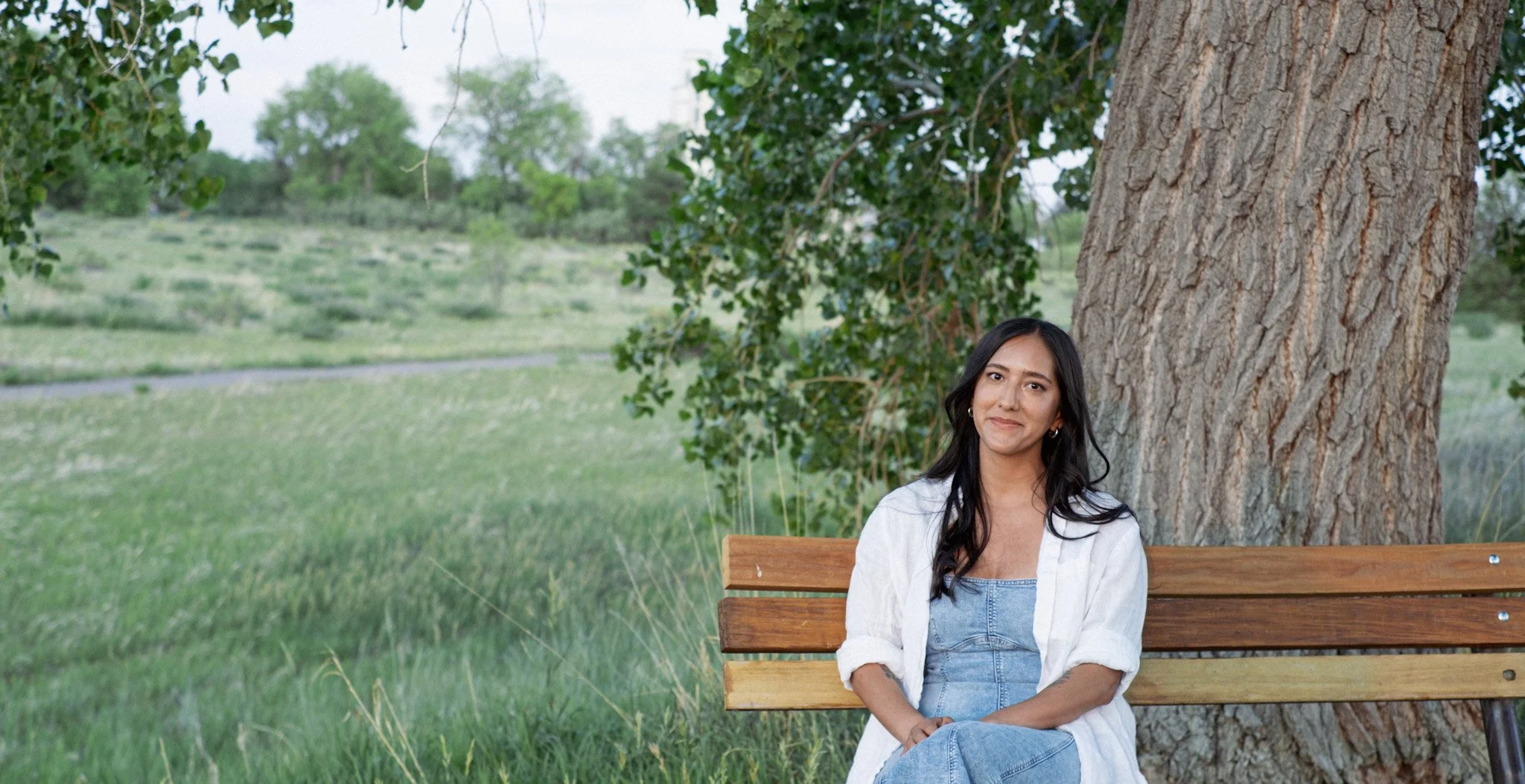 Photo of Dominique Guillen LCSW. She has long dark hair, a warm brown completion and is looking in to the camera and smiling with denim dress and linen button up on in front of a green meadow framed by a large sturdy tree.