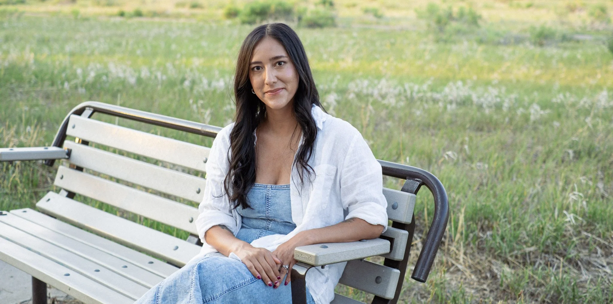 Photo of Dominique Guillen LCSW. She has long dark hair, a warm brown completion, wearing a white linen button up over a denim dress, and is looking in to the camera and smiling. She's sitting on a bench in front of a green and yellow meadow