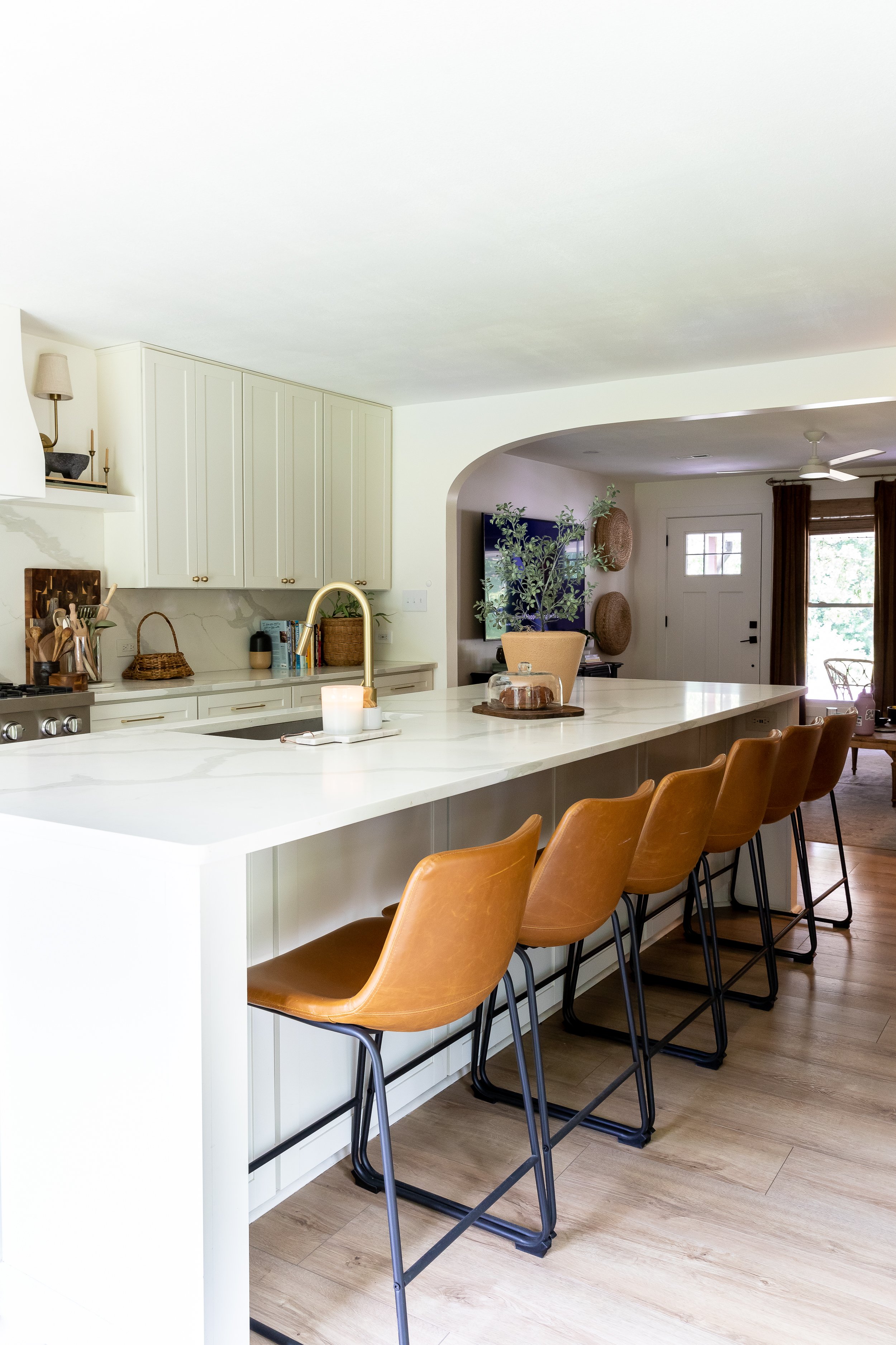 Modern kitchen with white cabinets, a large island, brown leather bar stools, and wood flooring. The island has a sink and is decorated with a candle and tray. Natural light filters in from a window in the adjoining room.