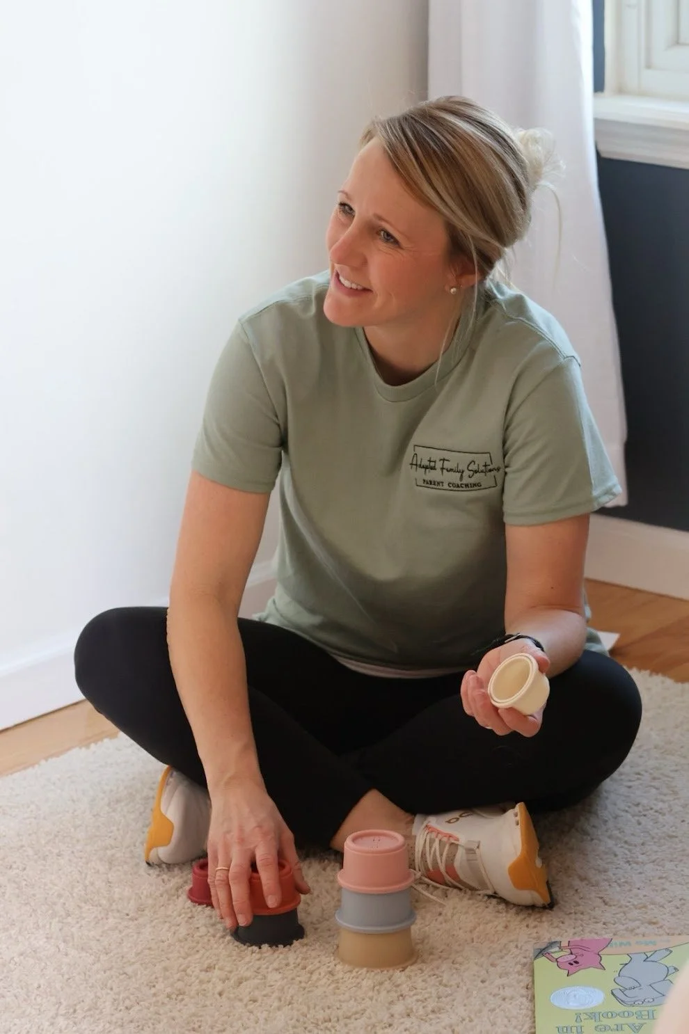 Woman sitting cross-legged on a rug, playing with stacking cups, smiling, wearing a gray T-shirt and athletic shoes, in a room with white walls and a window.