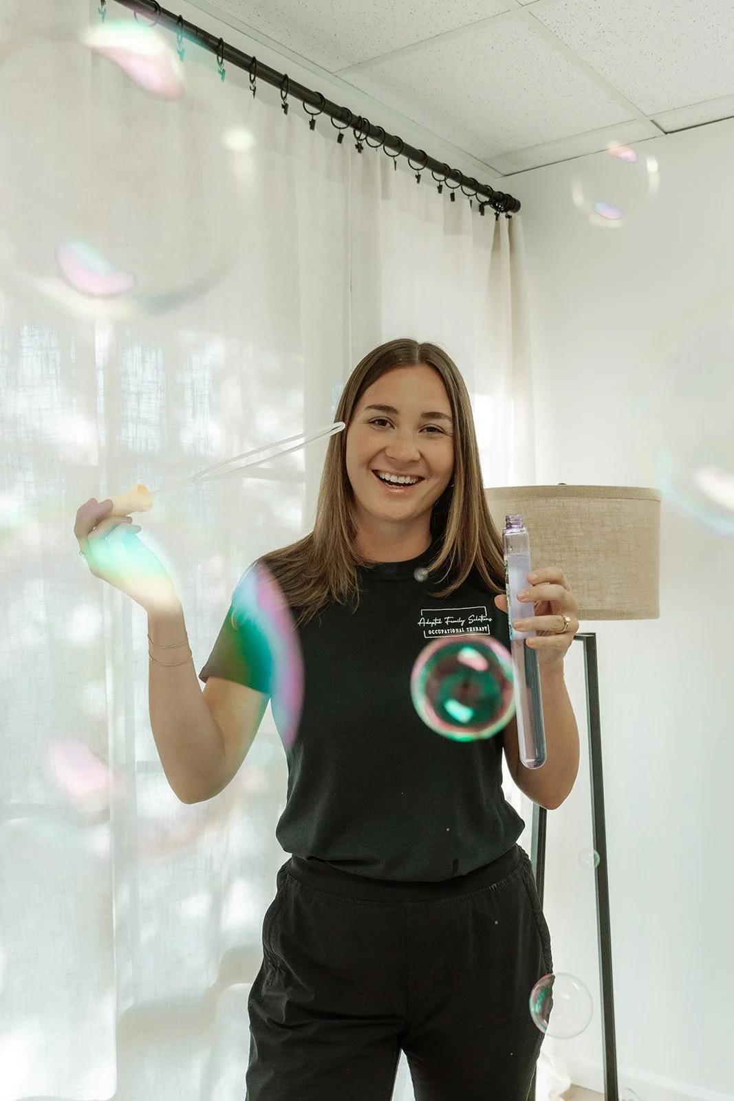 A woman smiling and holding a bubble wand with soap bubbles floating around her indoors.