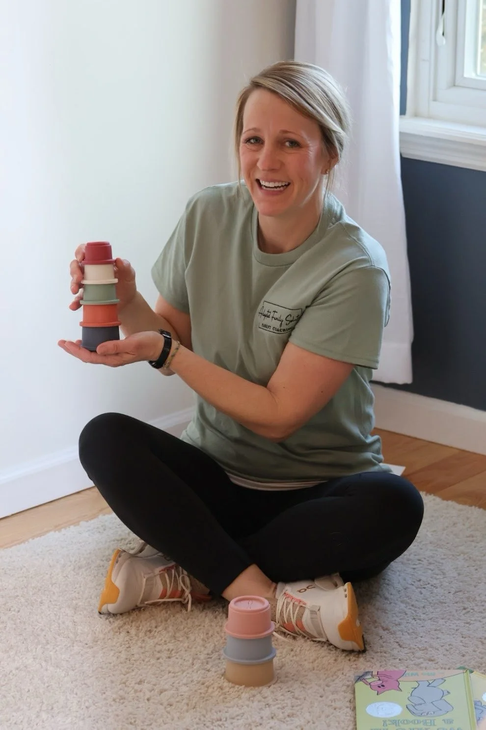 Woman sitting on a beige carpet, holding a stack of colorful cups and smiling at the camera. There is a small stack of cups on the floor and a partially open book nearby. She is in a bright room with white curtains, a window, and dark blue wall.