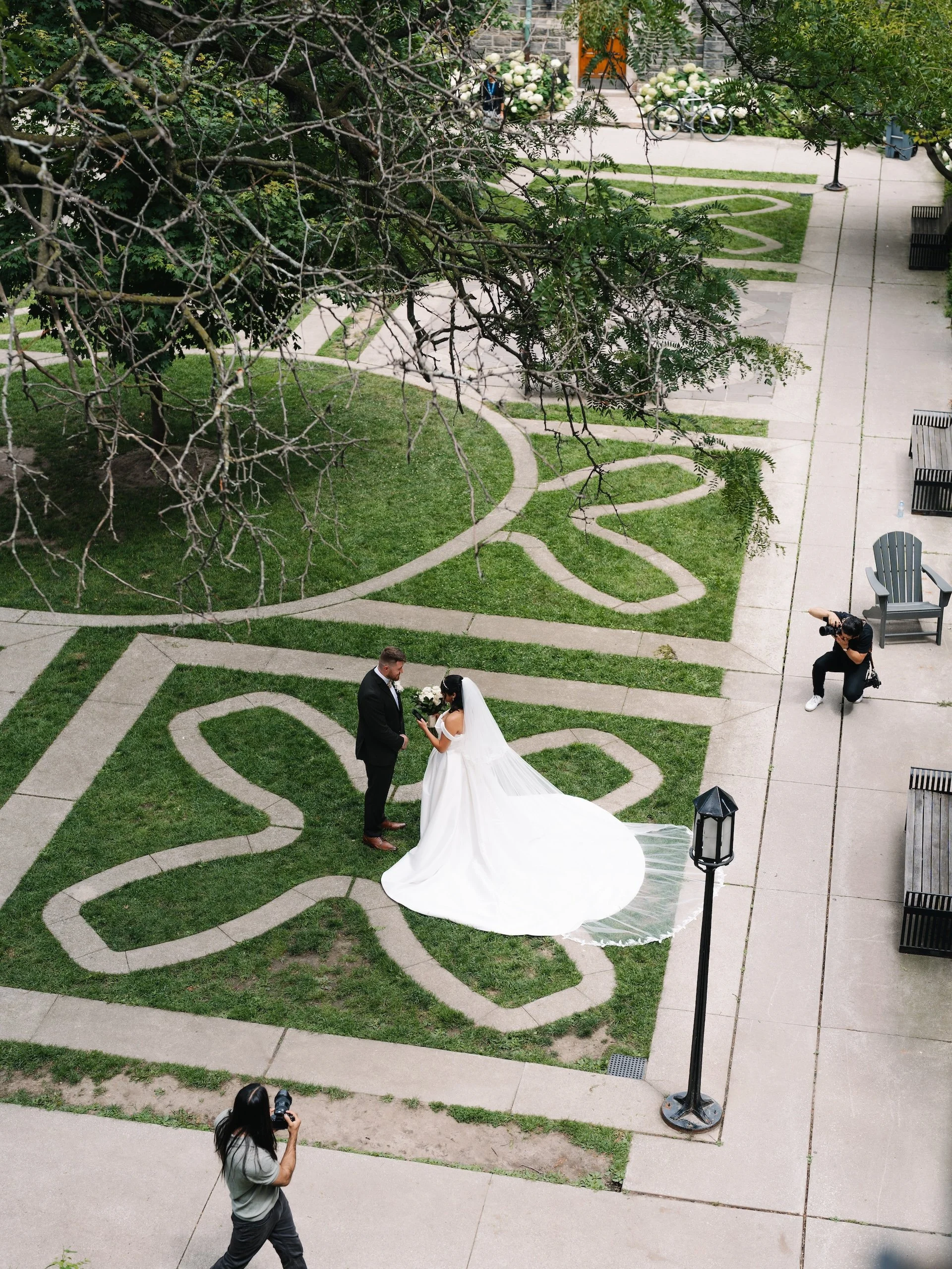 Wedding couple standing on a decorative garden path, with photographers capturing the moment, in an outdoor park setting.