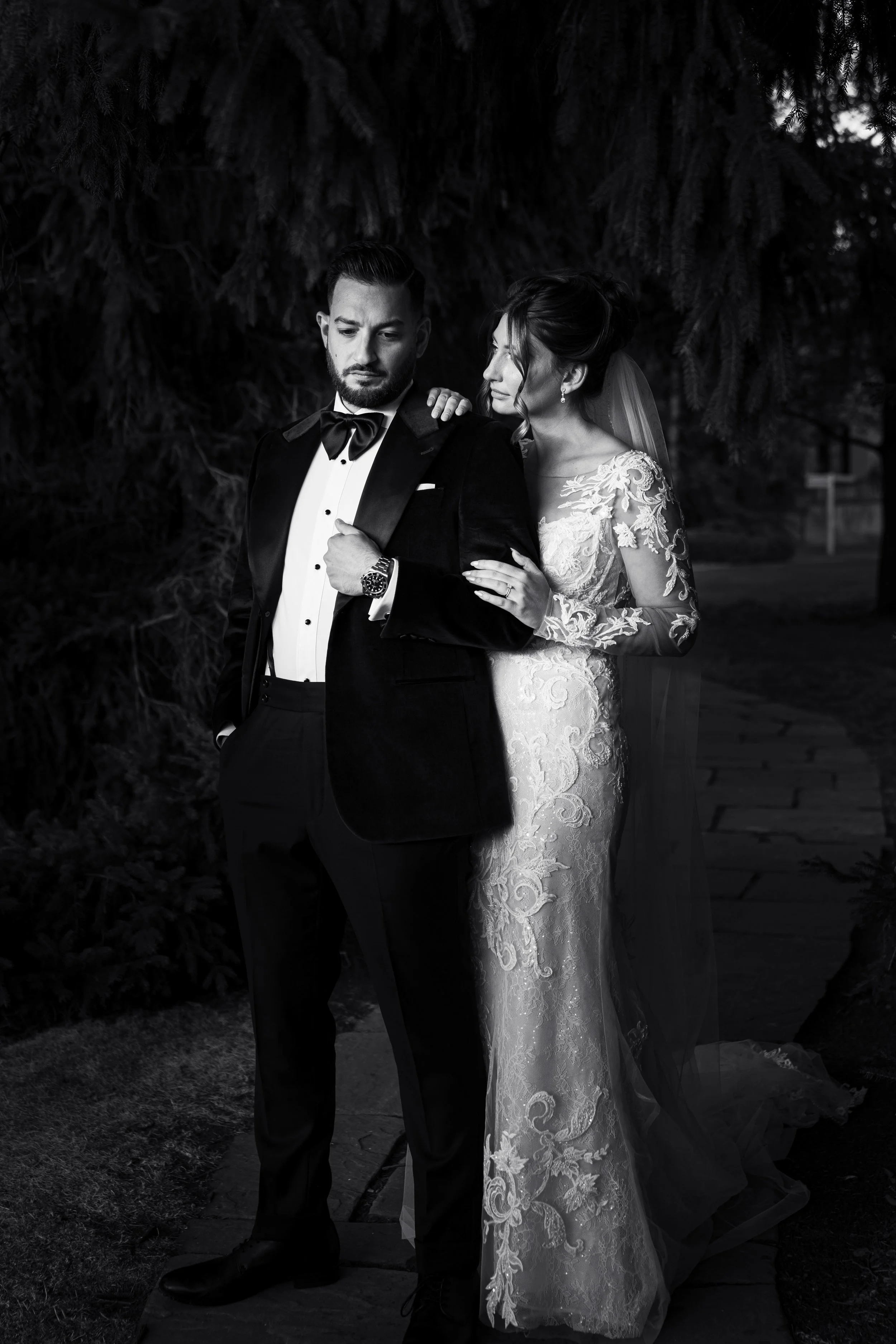 Black and white photo of a bride and groom outdoors. The groom is dressed in a tuxedo and bowtie, with one hand in his pocket, looking down. The bride in a lace wedding gown is leaning on his shoulder, with her hand on his arm, and wears a veil. They