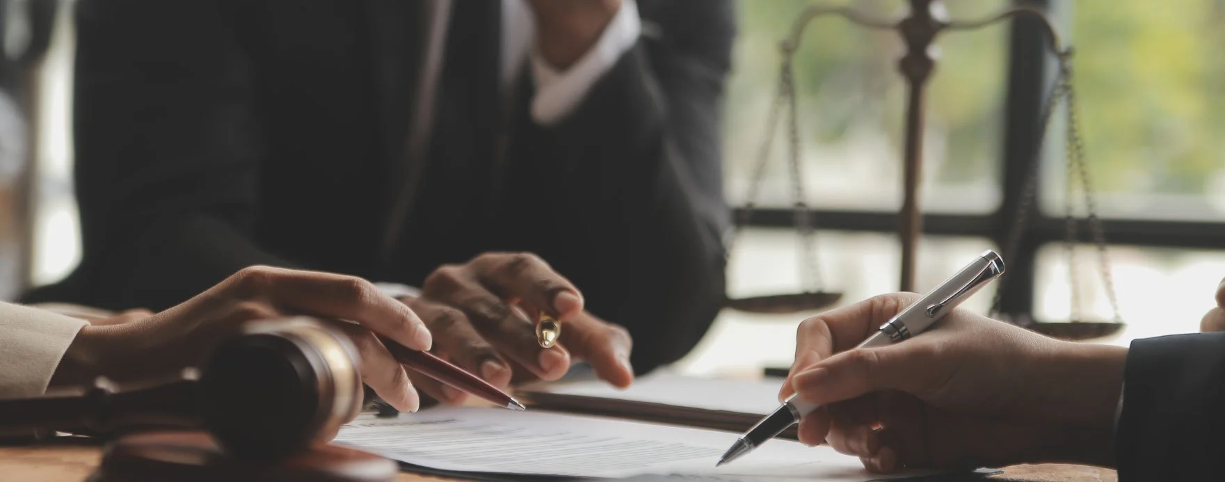 Two people in formal attire signing documents at a table in a legal office, with legal scales visible in the background.