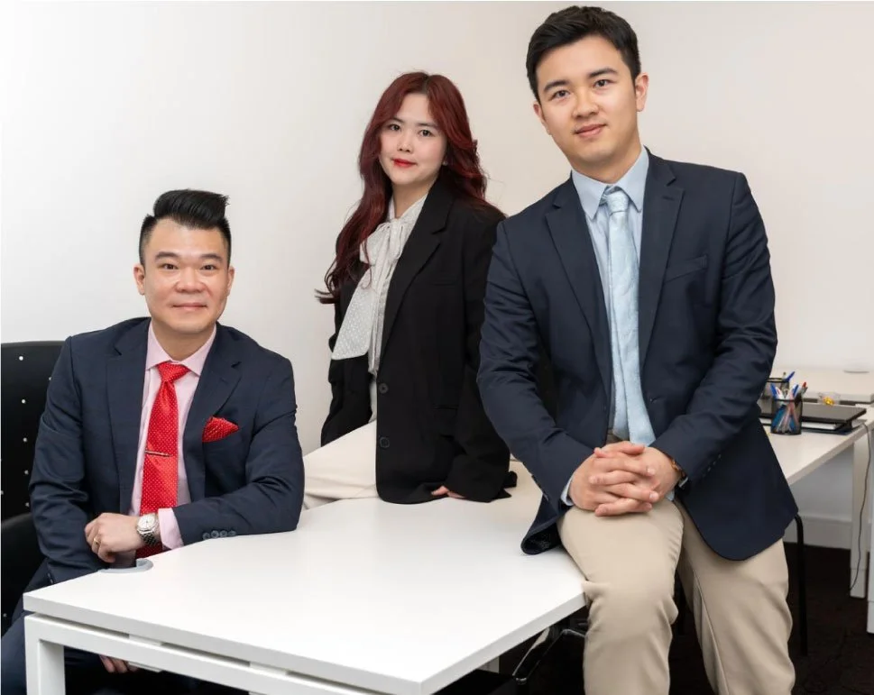Three young professionals in business attire in an office, two men seated at a desk and a woman standing behind them.