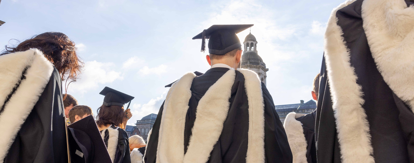 Graduates wearing caps and gowns with white stoles gather outdoors under a partly cloudy sky, with a historical building in the background.
